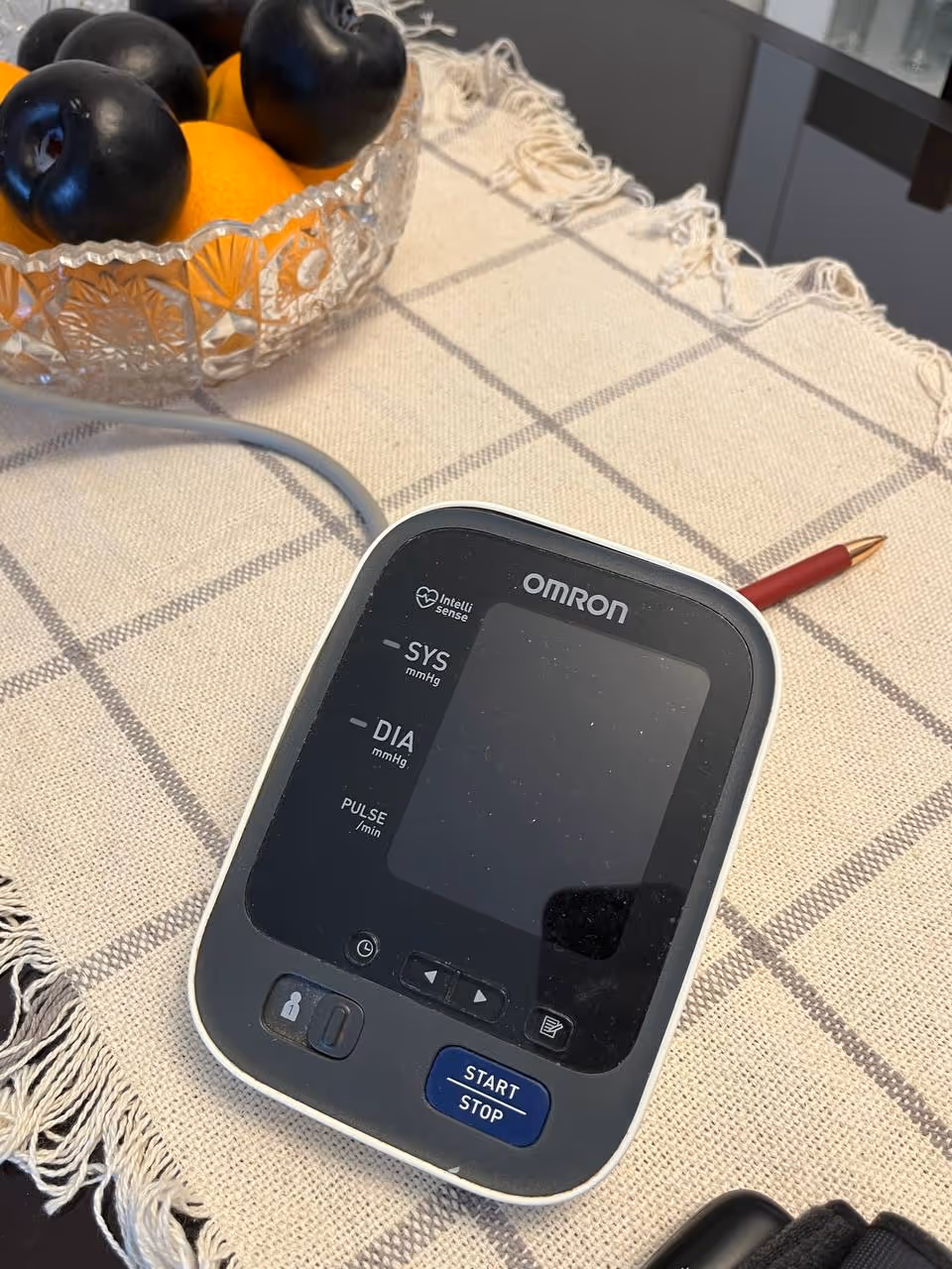 Omron blood pressure monitor on a checkered tablecloth beside a fruit bowl in a home setting.