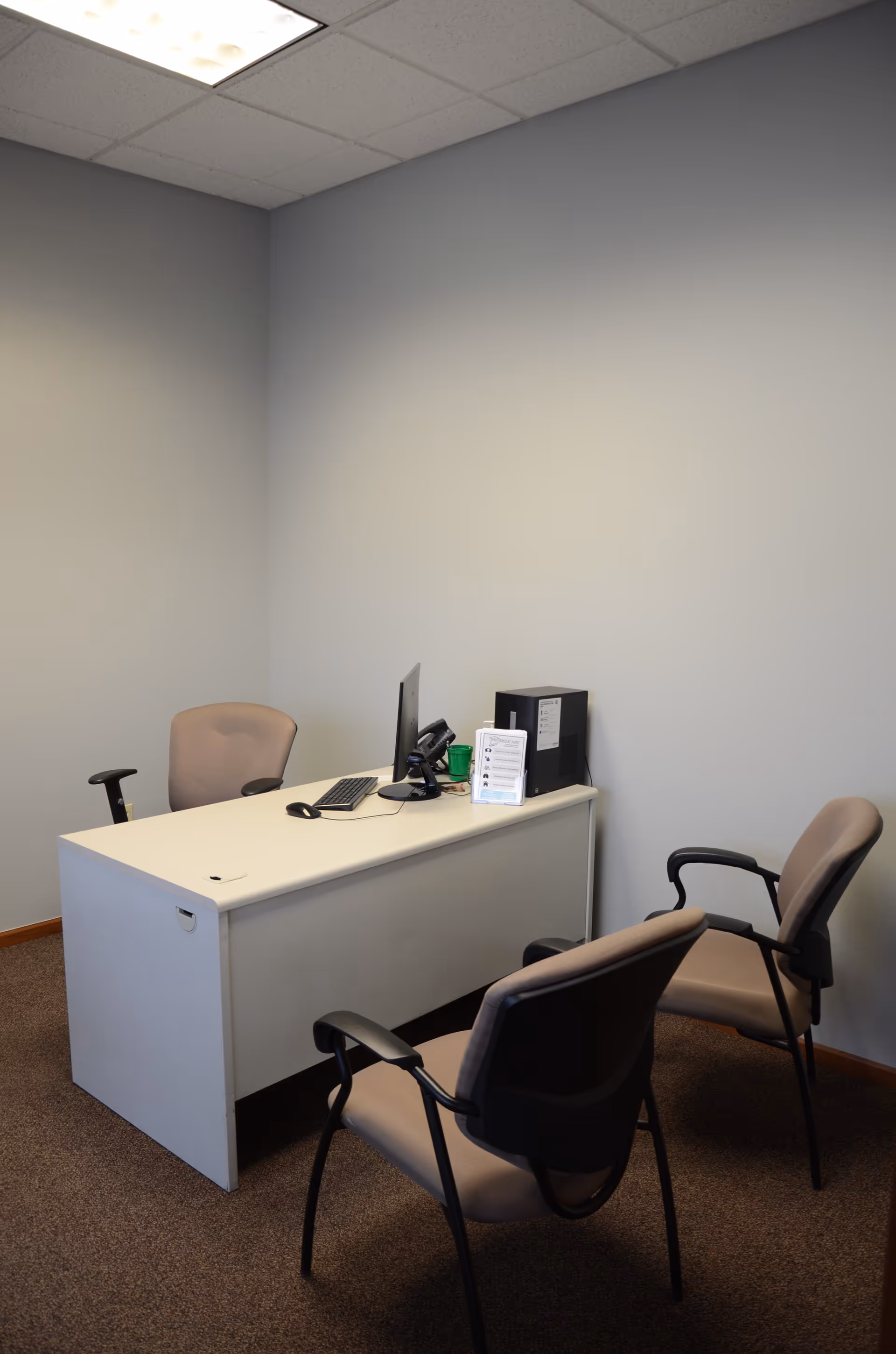 Small office with a white desk, computer, phone, and three chairs against pale walls.