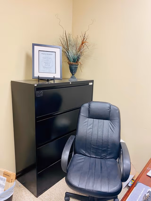 Office corner with filing cabinet, framed document, decorative vase, and black swivel chair.