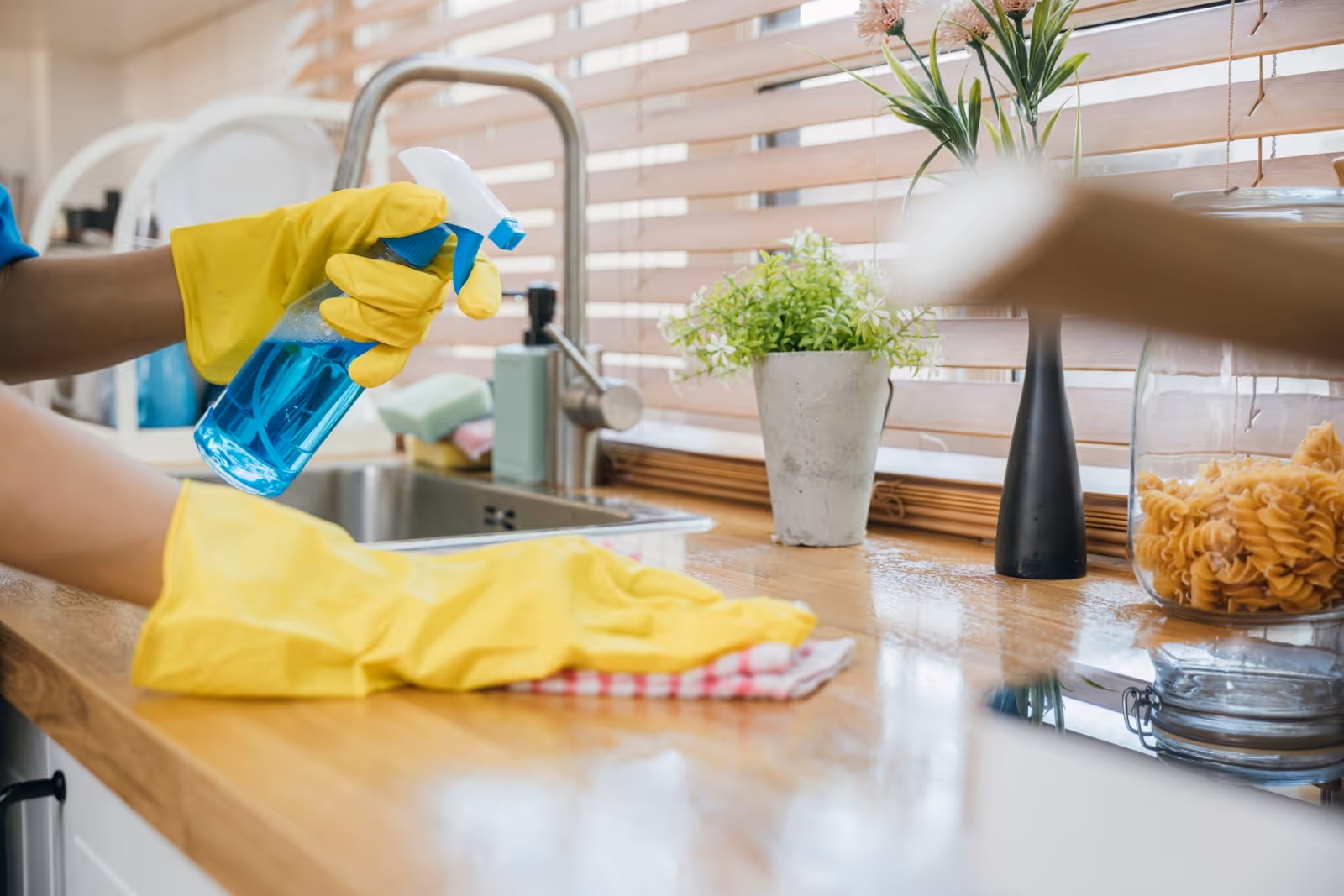 Yellow-gloved hands spray cleaner on a kitchen counter beside a sink and plants.