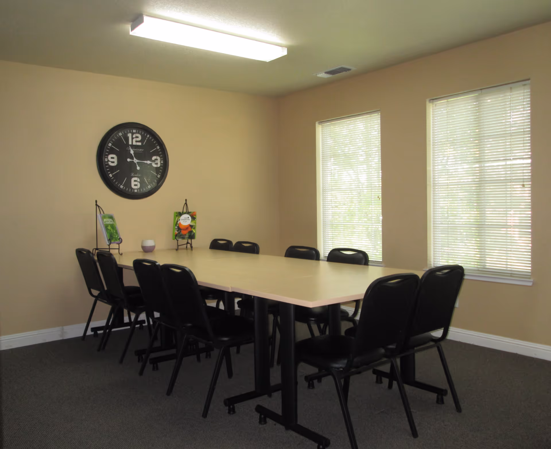 A beige interior meeting room with a long table, black chairs, clock, and large windows.