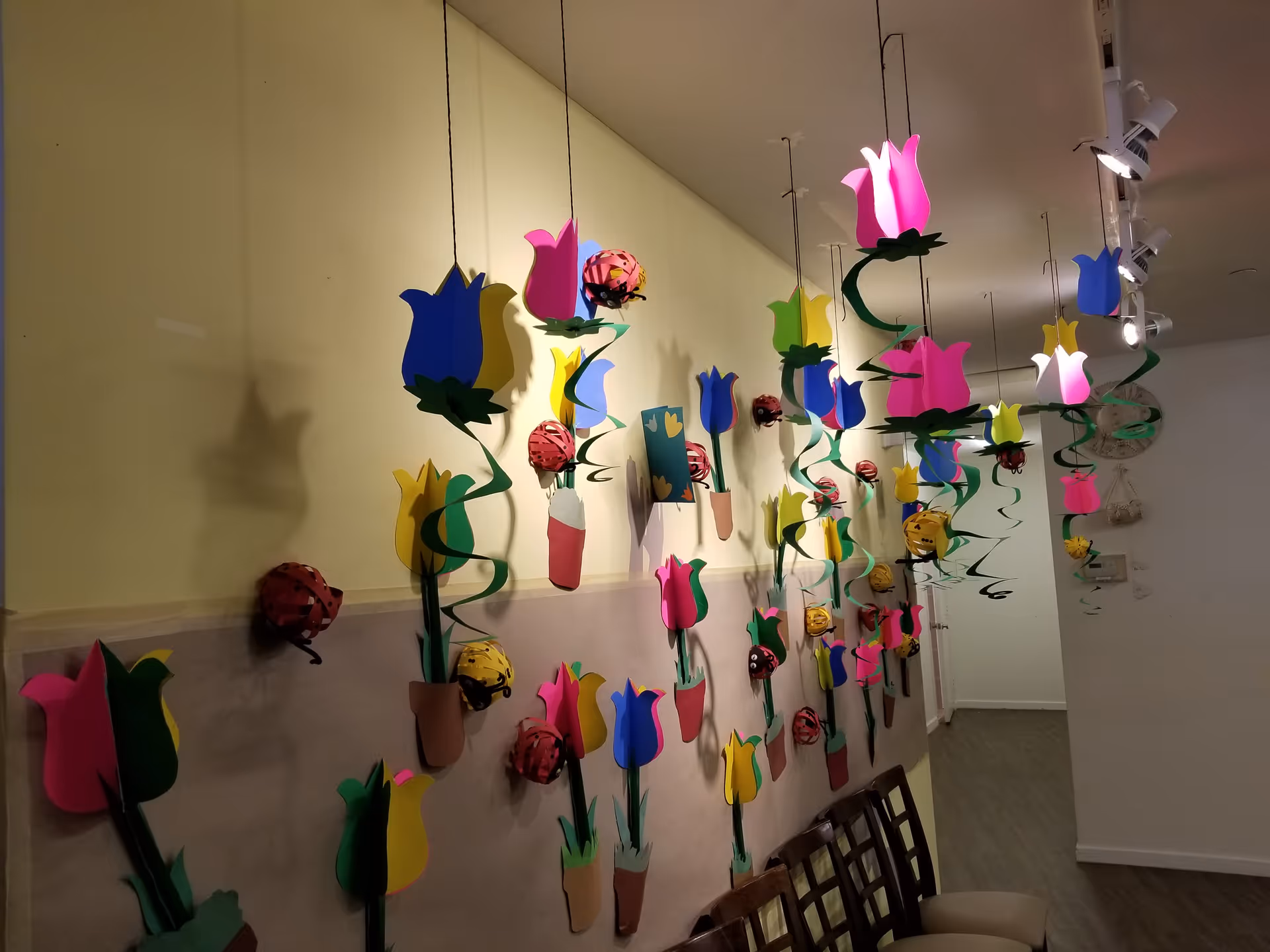 Colorful paper flowers hang from the ceiling along a hallway with chairs.