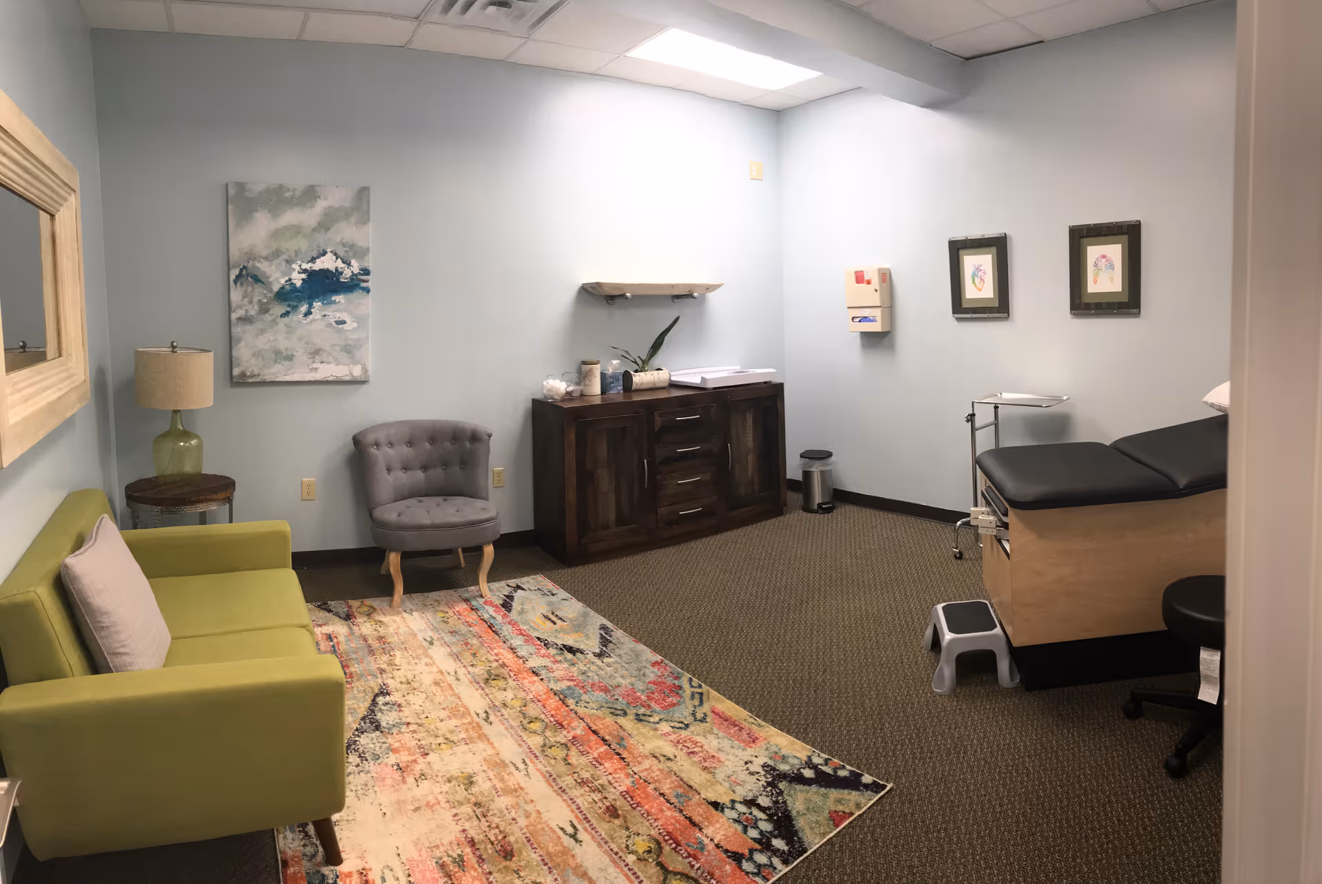 A cheerful waiting area with green sofa, armchair, artwork, and a medical exam bed.