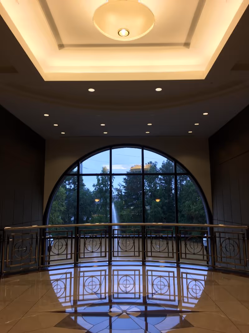Spacious lobby with large arched window, outside fountain and trees, polished tiled floor.