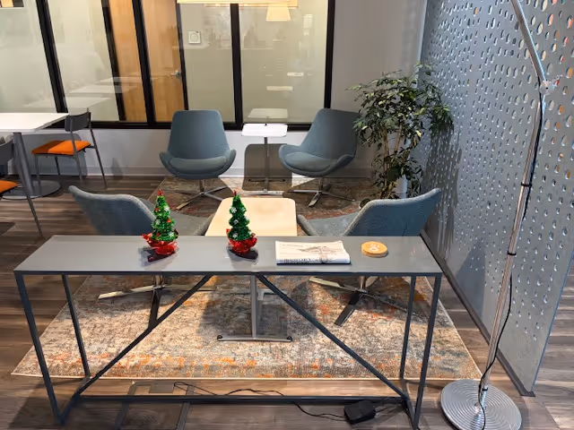 Modern indoor lounge with a desk, multiple chairs, rug, plant, and small Christmas trees.