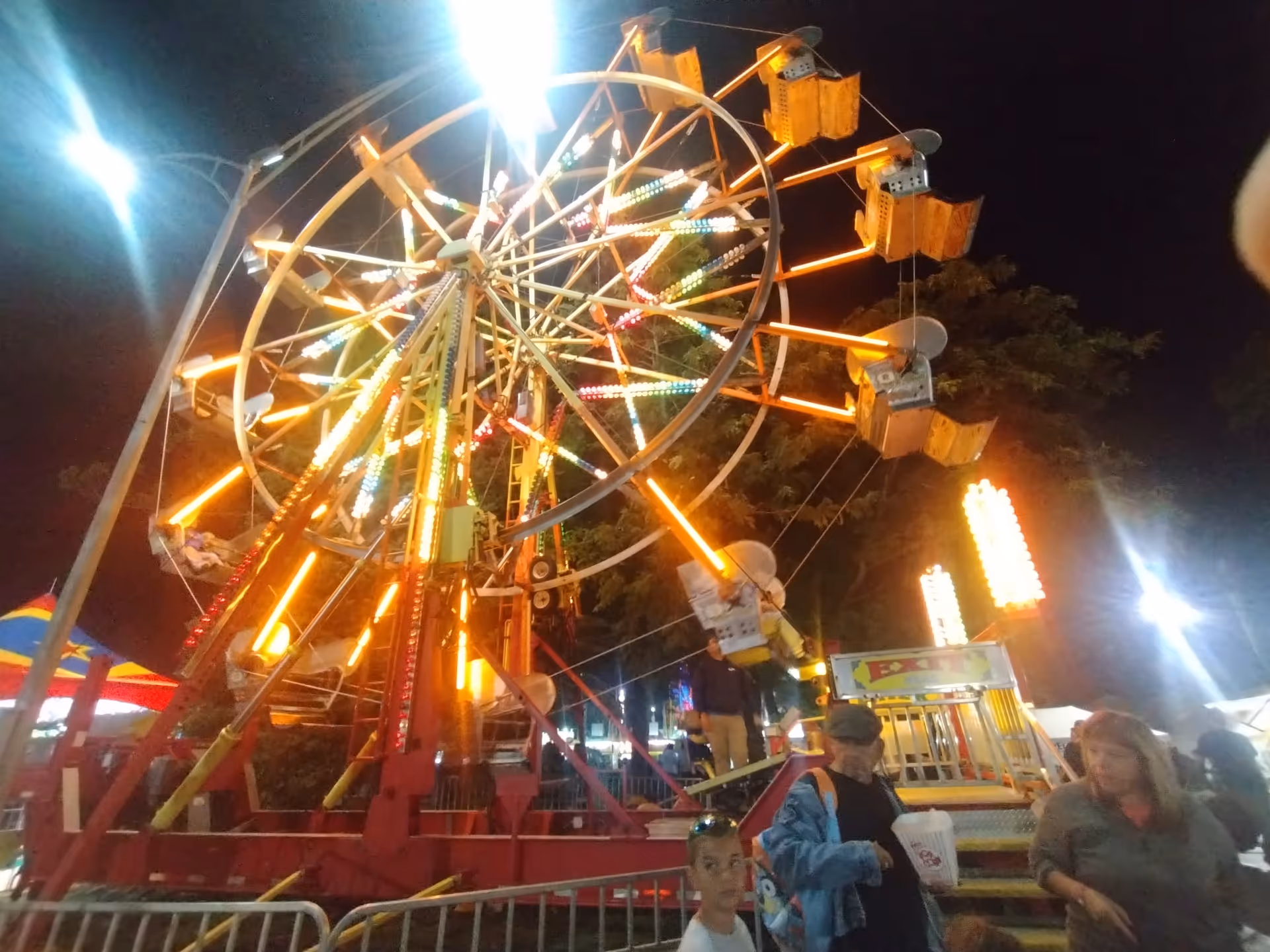 Nighttime carnival scene featuring a brightly lit Ferris wheel and people nearby.