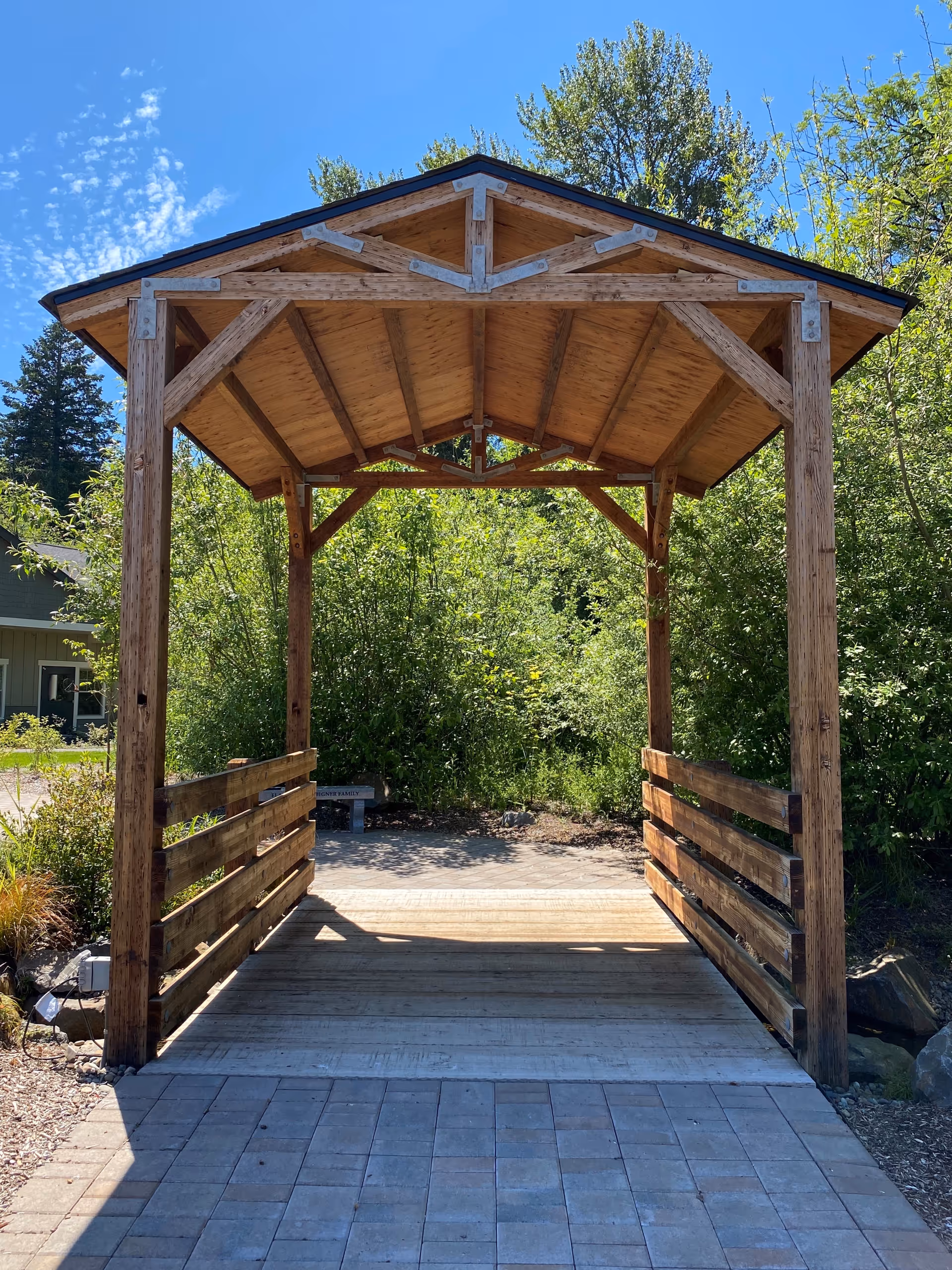 Wooden covered pavilion over a brick-paved path in a garden setting.