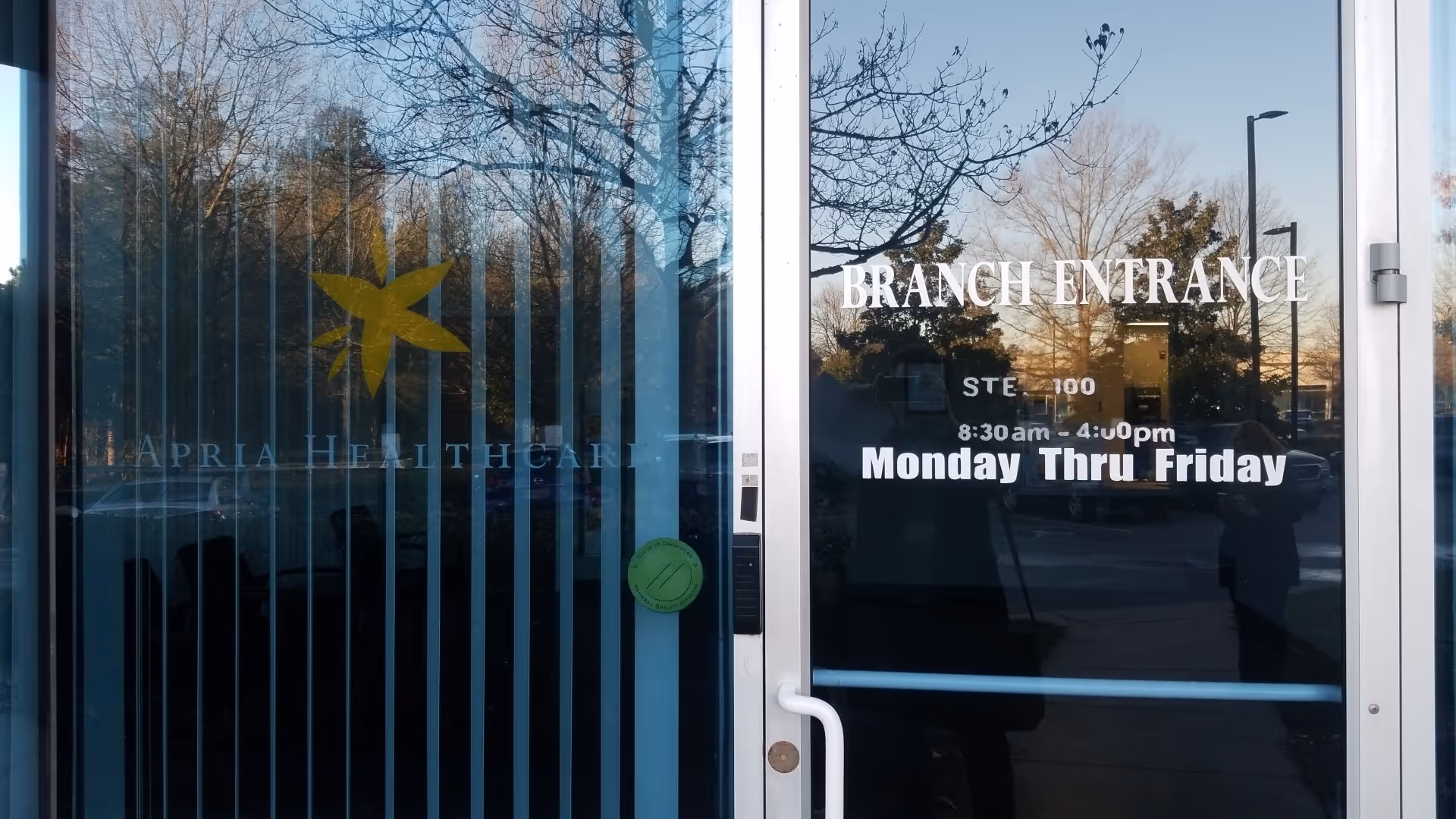 Glass doors show Apria Healthcare branding and 'Branch Entrance' with reflections of trees outside.