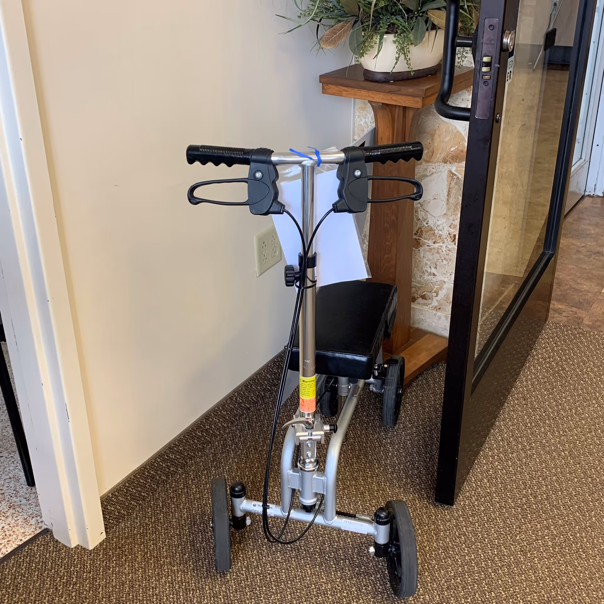 A four-wheeled mobility aid with handlebars and a seat in a hallway near a glass door.