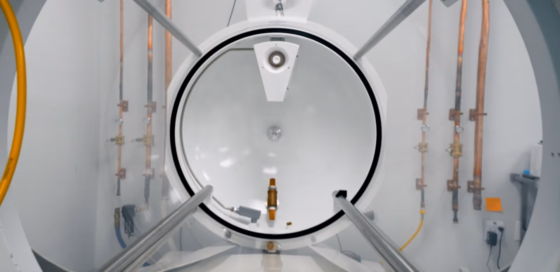 Inside a circular hyperbaric chamber, with copper pipes and support rails surrounding.