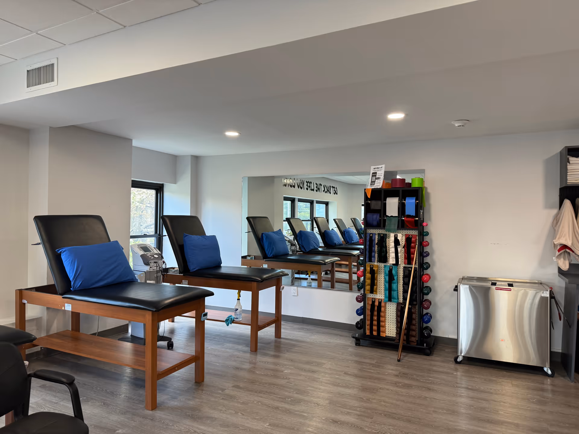 Bright physical therapy treatment room with adjustable chairs, a mirror, and equipment rack.