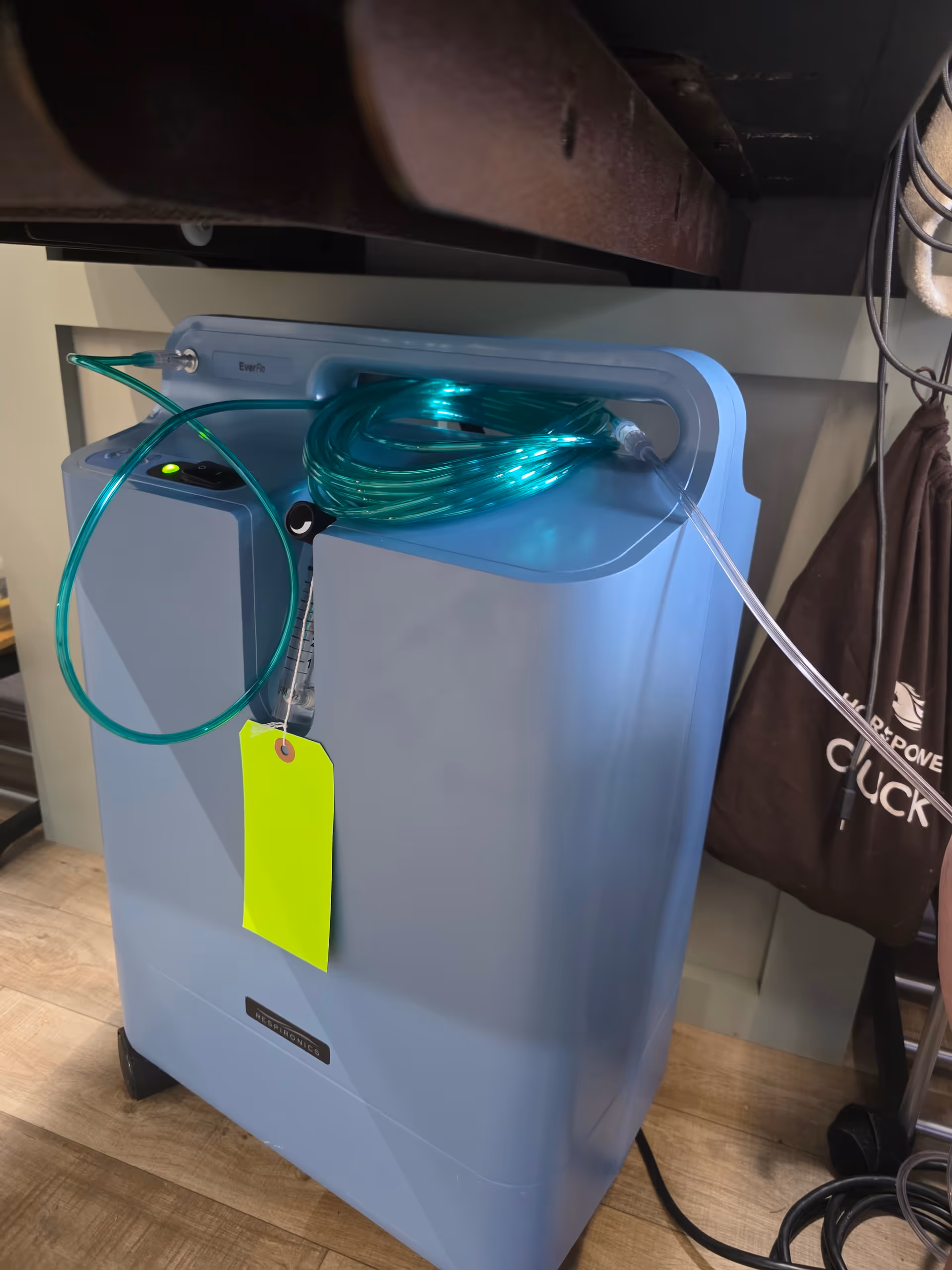 Blue oxygen concentrator with tubing and a neon tag indoors on a wooden floor.