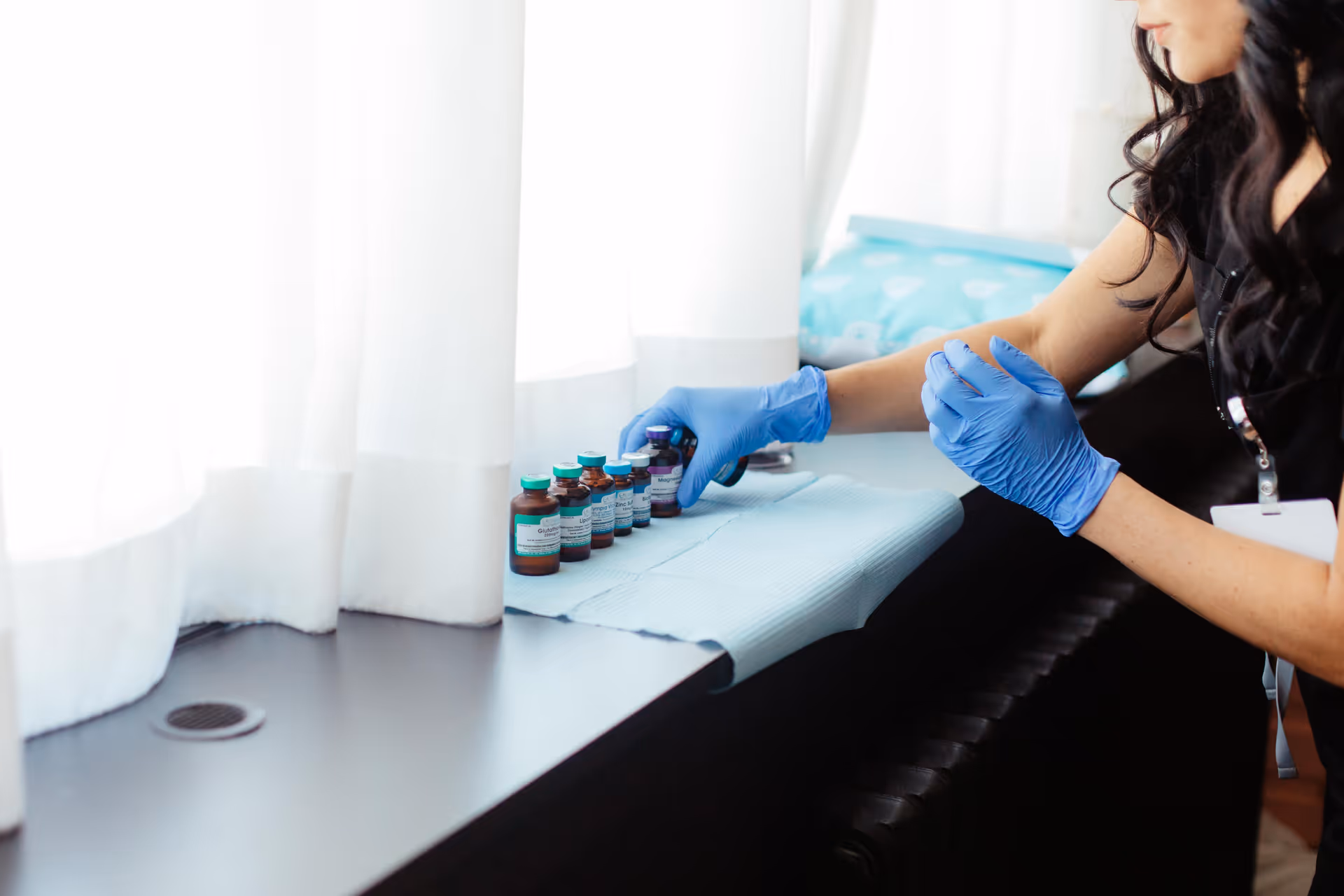 Gloved healthcare worker arranges small medication vials on a windowsill beside white curtains.