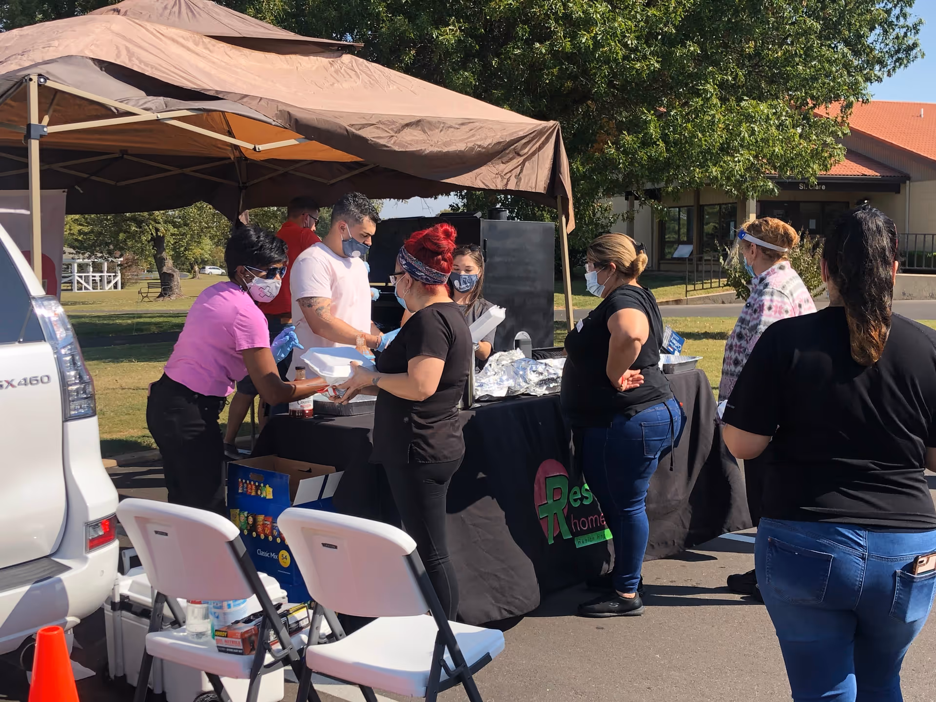 A group of people wearing masks gather around a food table under a brown canopy outdoors.