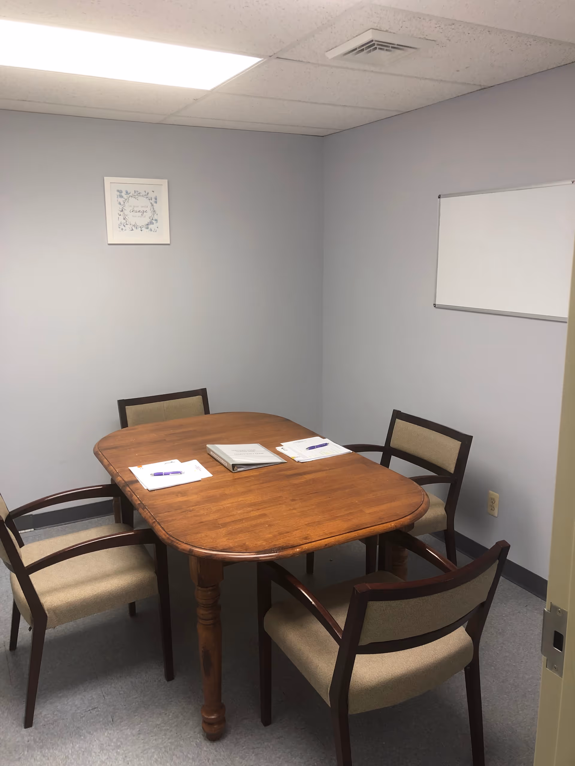 A small conference room with a wooden table, four chairs, papers, and a whiteboard.