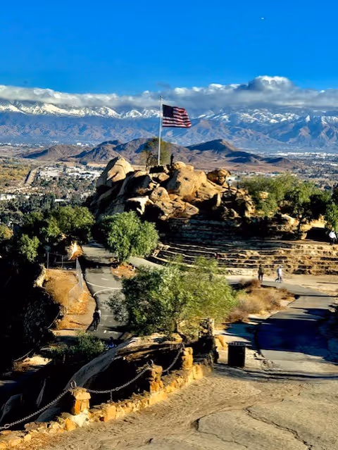 American flag atop a rocky overlook with trees and snow-capped mountains in the distance.