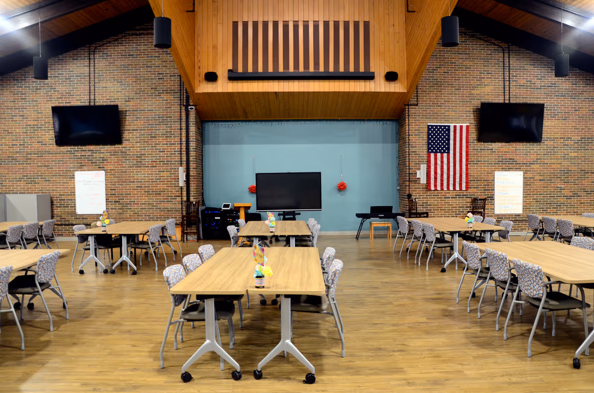 A spacious multipurpose dining hall with multiple tables, chairs, brick walls, and an American flag.