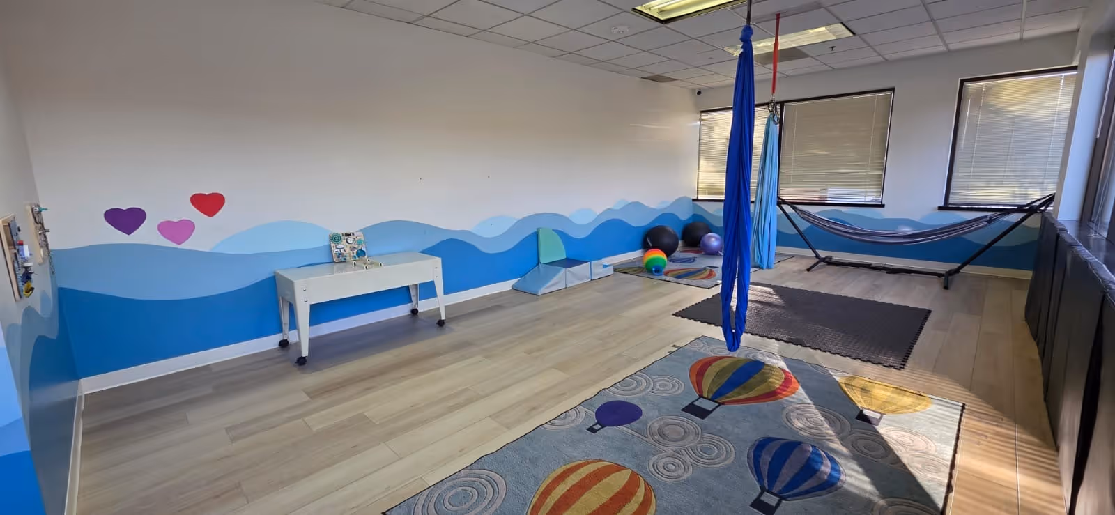 Bright therapy room with aerial fabrics, mats, and a blue wavy wall mural.