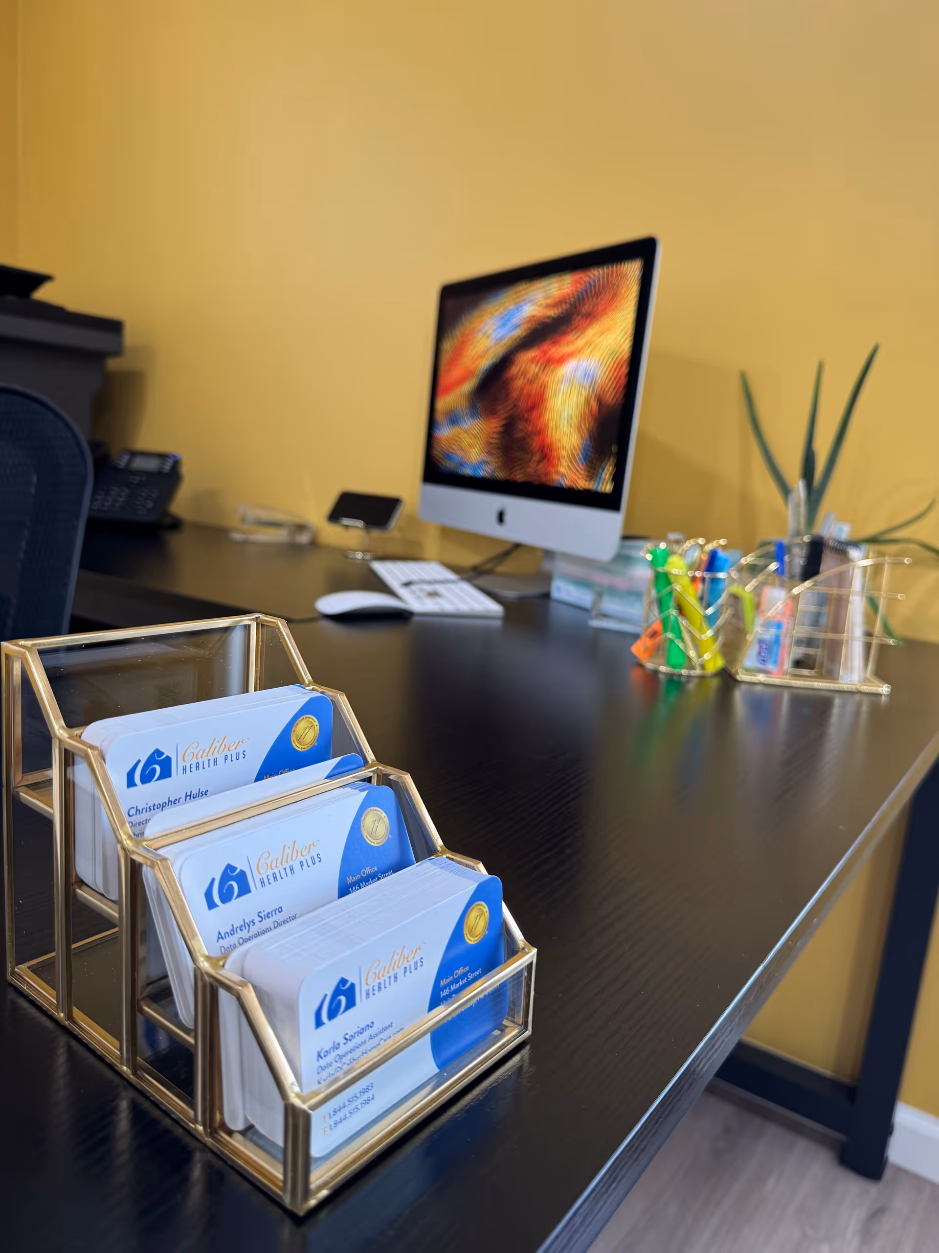 Office desk with an iMac monitor and a gold Caliber Health Plus card holder.