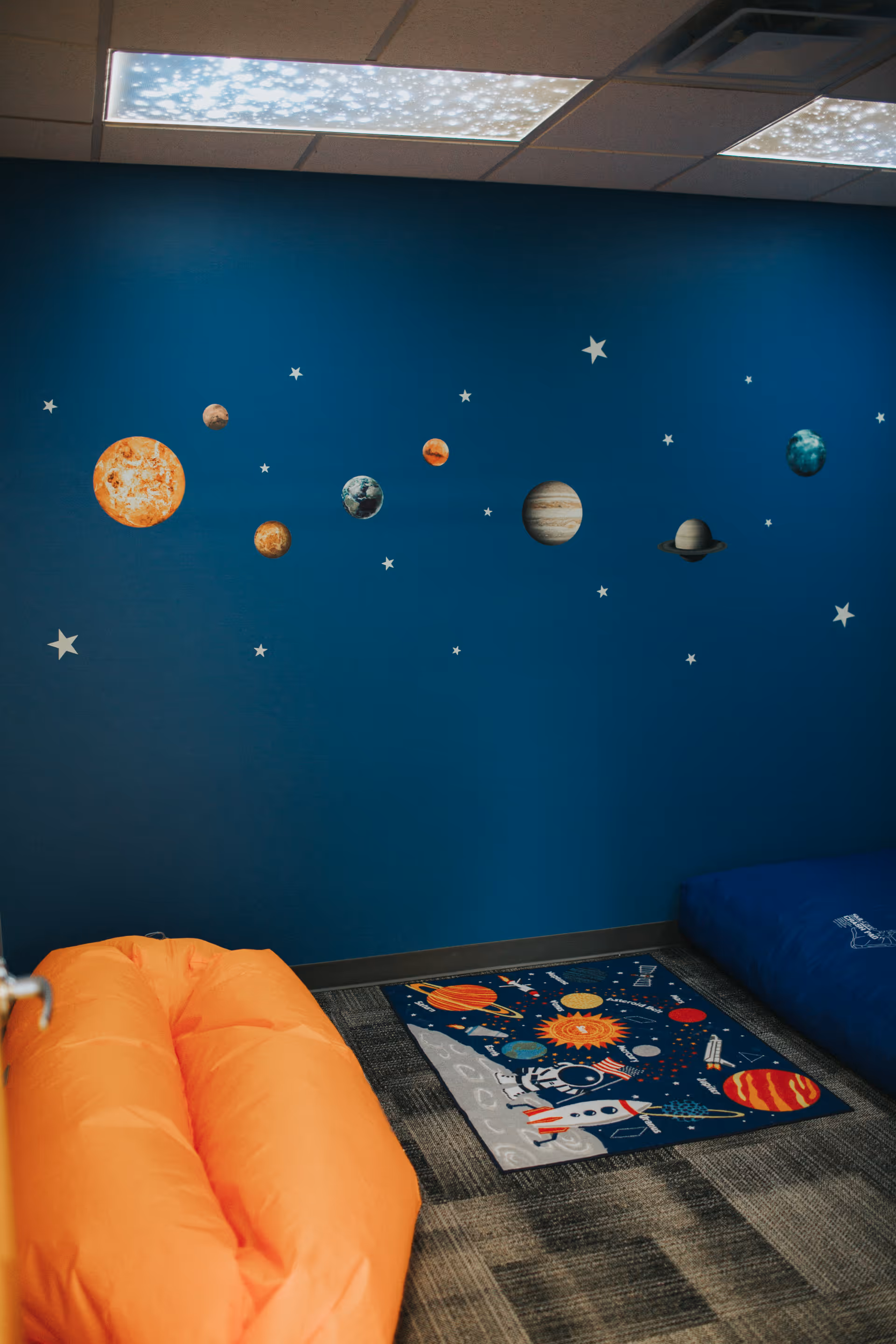 Blue wall with space-themed decals, rug, and orange bean bag chairs in a therapy room.
