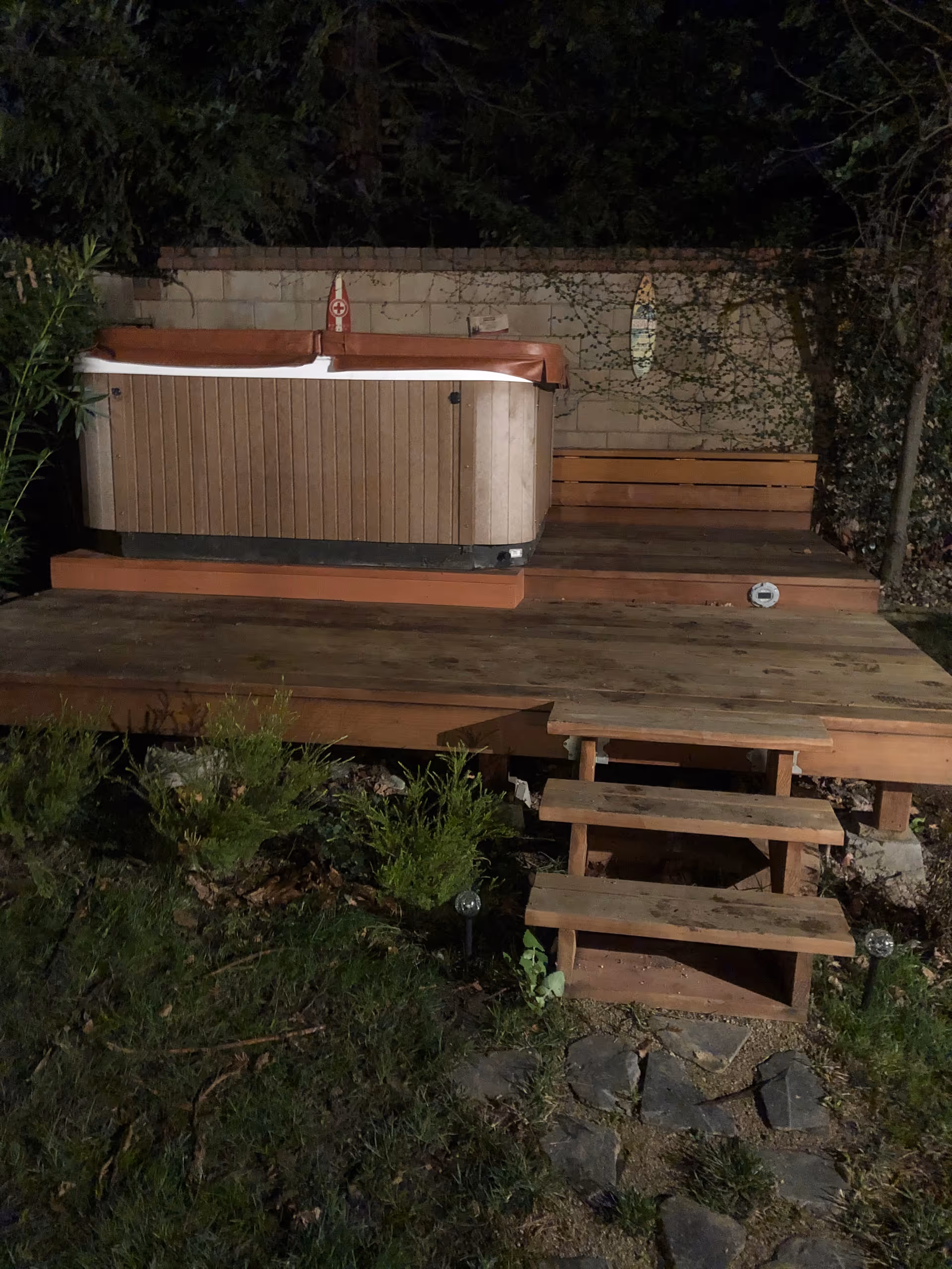 Nighttime outdoor deck with a hot tub, wooden steps, and surrounding greenery.
