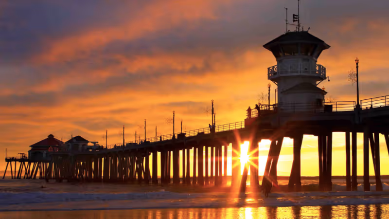 Sunset over a long pier with a tower, calm water reflecting warm orange sky.