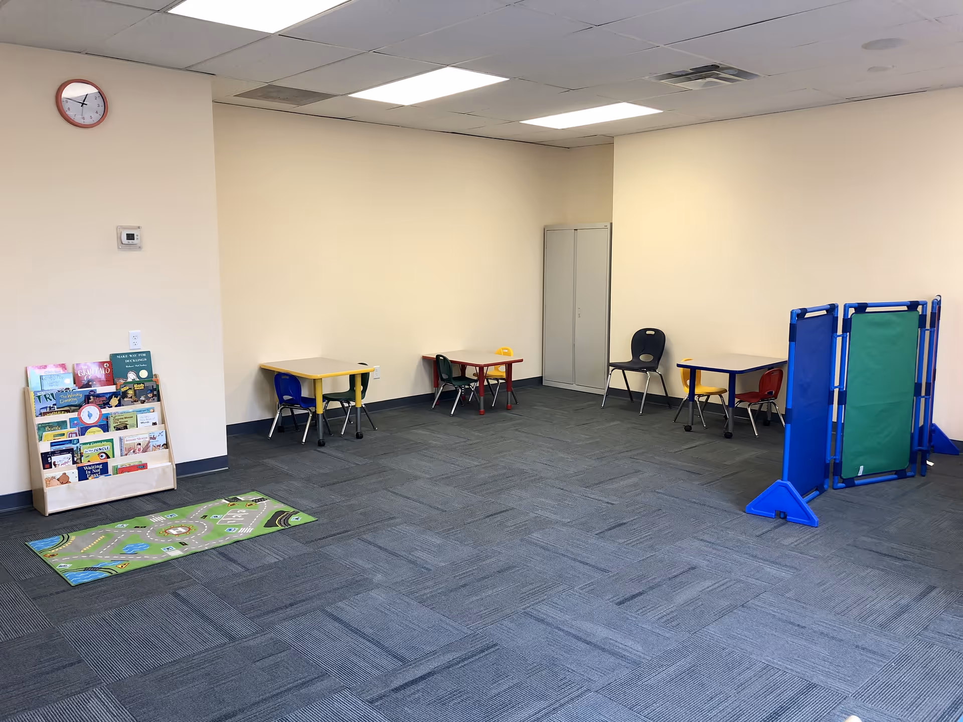 An empty activity room with small tables, chairs, a book rack, mats, and a divider.