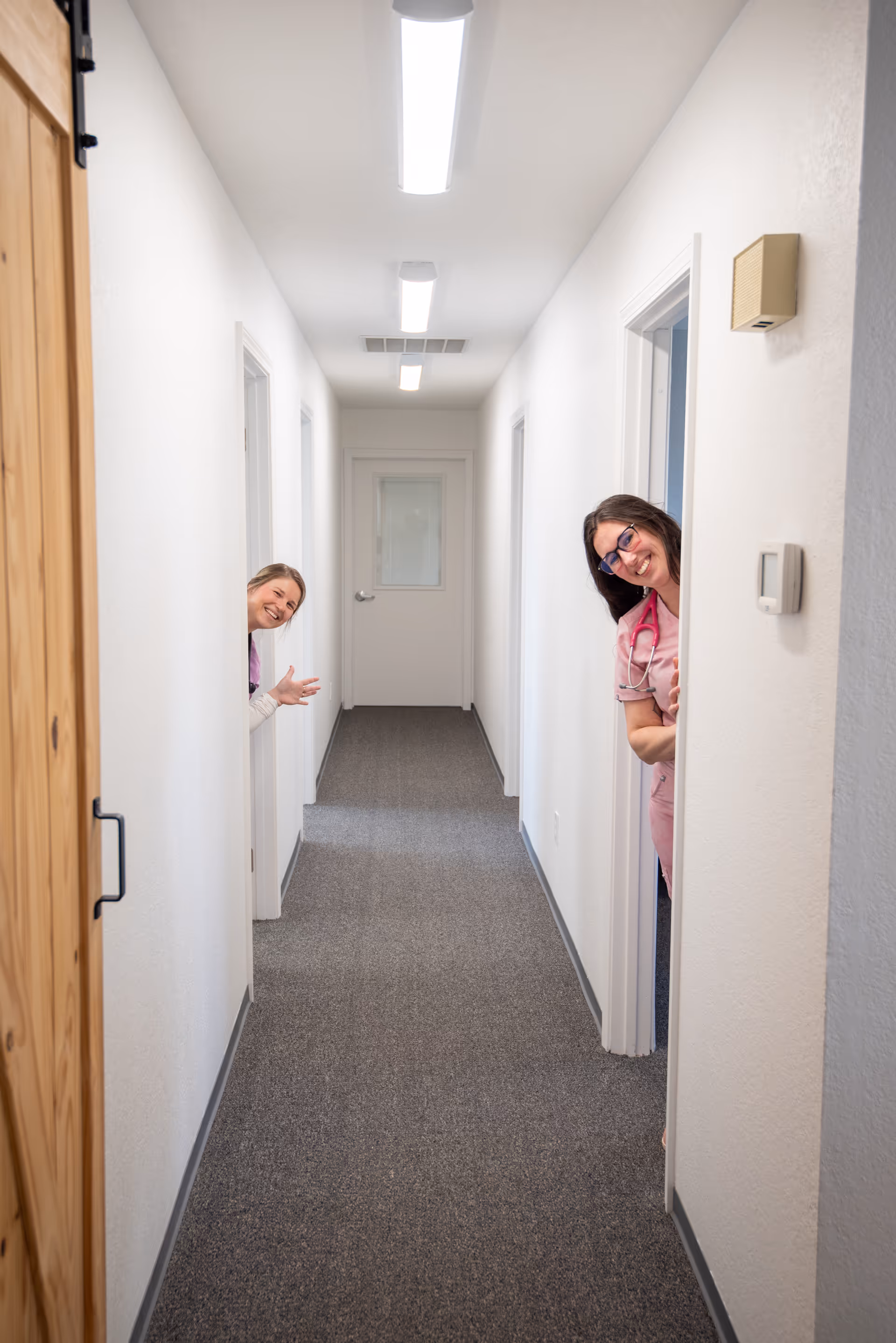Two staff members peek from doorways along a bright hallway.