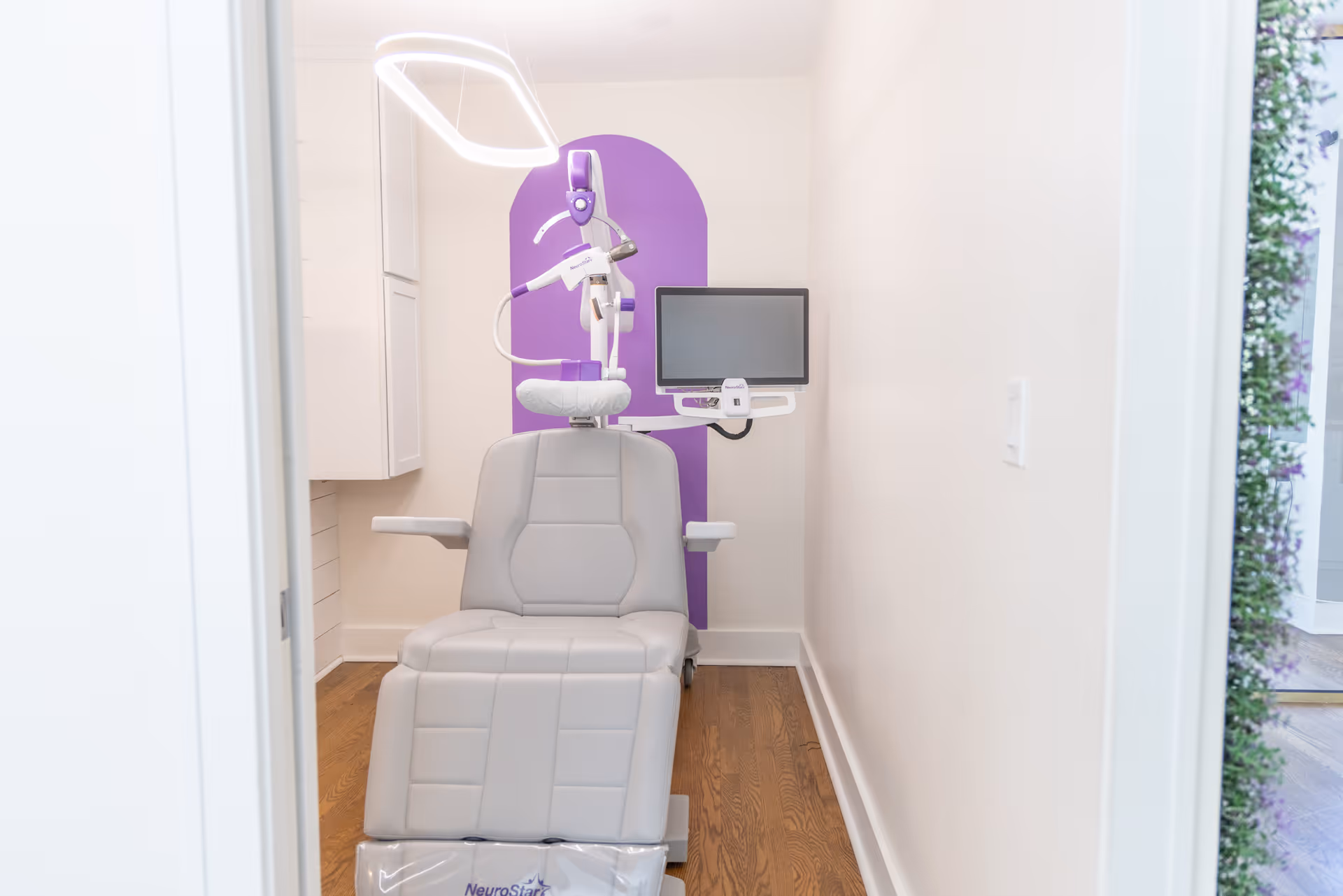 Bright interior treatment room with a reclining chair, monitor, and purple backdrop.
