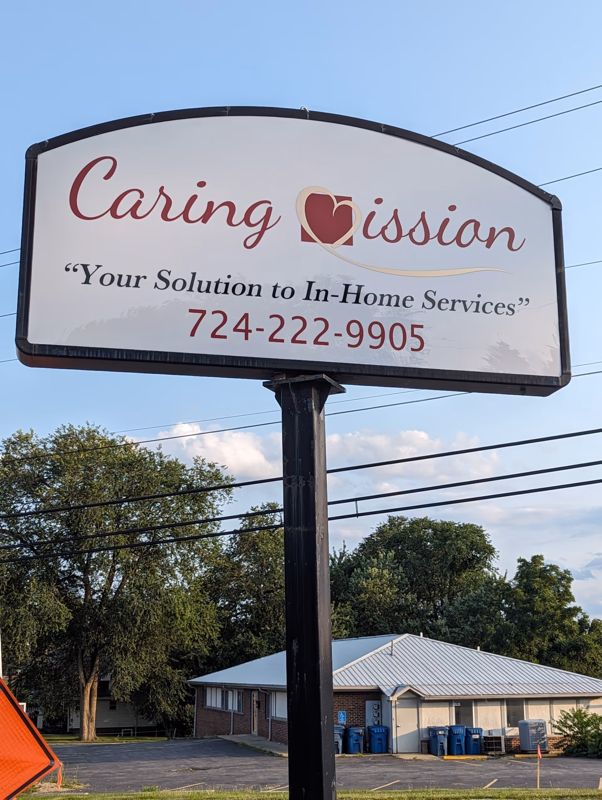 Large Caring Mission sign on a post with phone number, outdoor building behind.