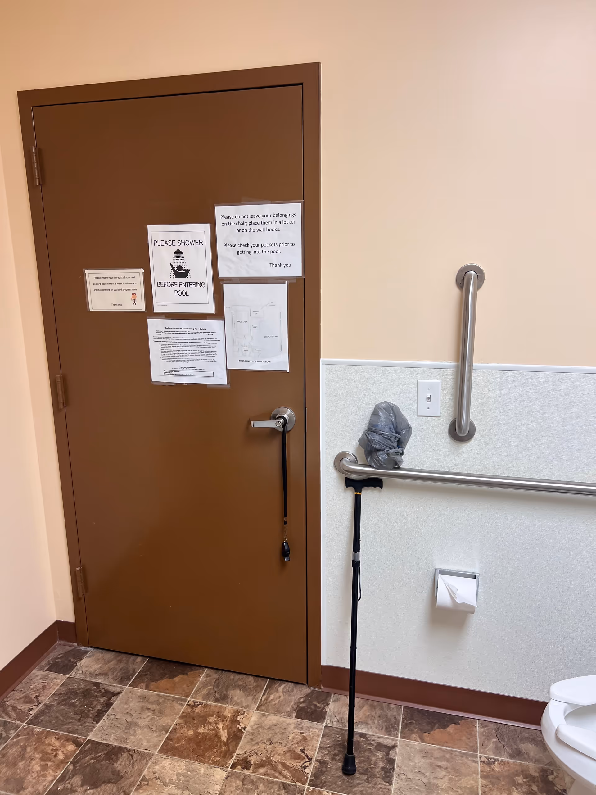 Brown door with notices beside an accessibility rail, cane, and toilet paper.
