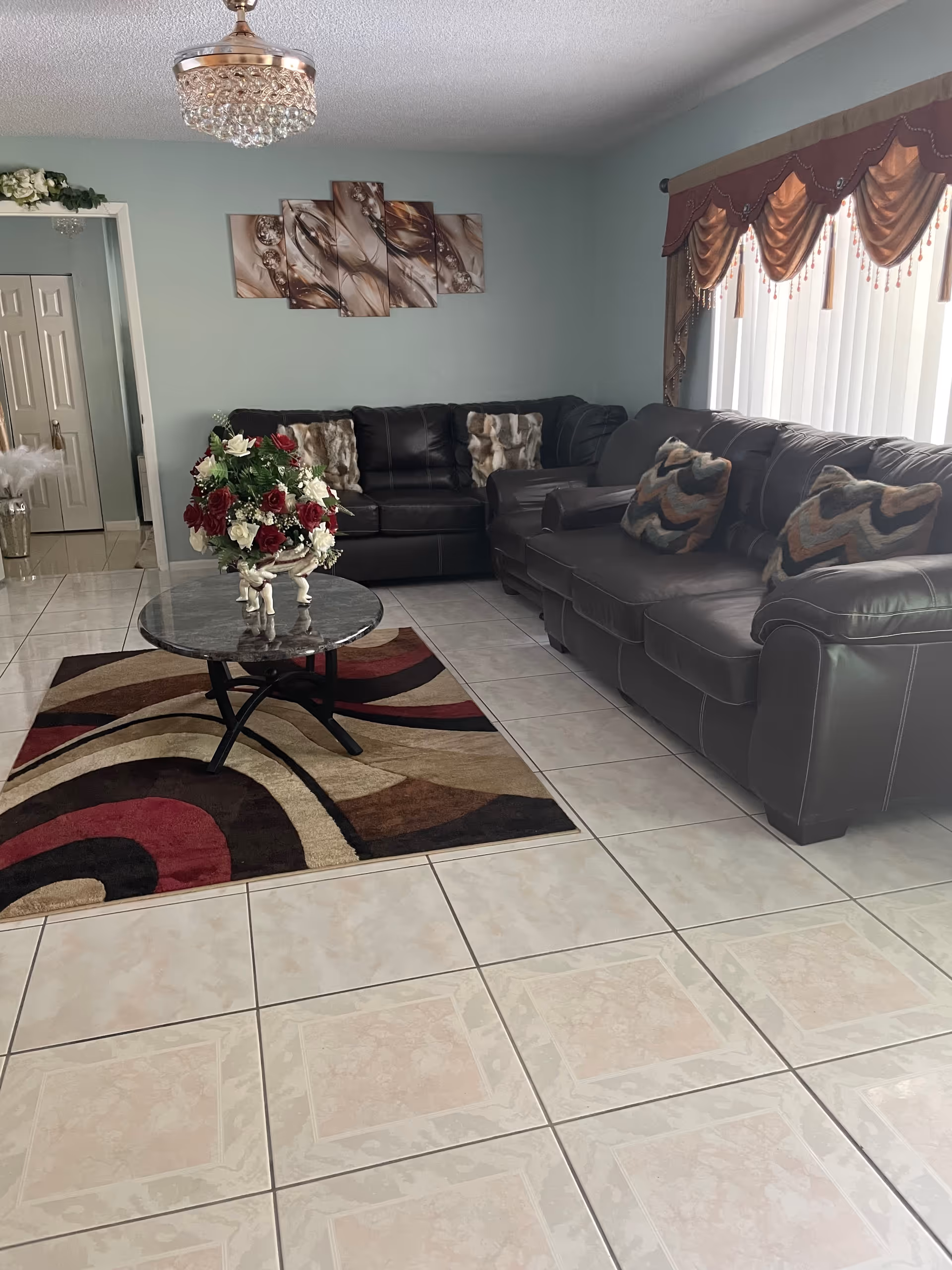 Common living room with leather sofas, glass coffee table, rug, chandelier, and draped windows.