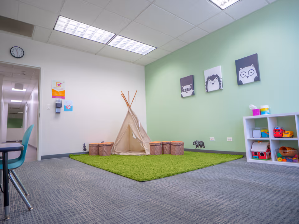 Interior playroom with a teepee tent, green carpet, shelves of toys, and wall art