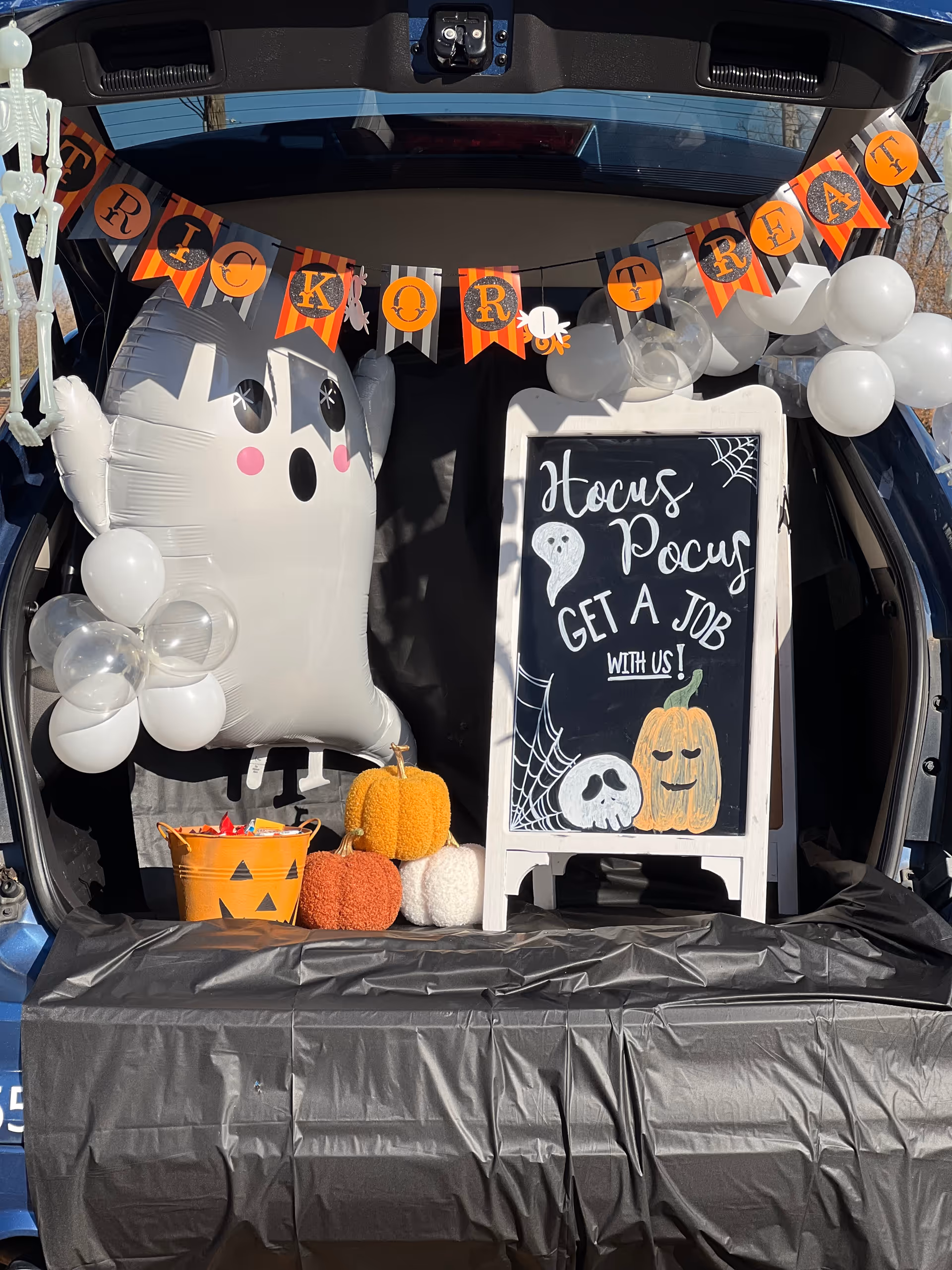 Open car trunk decorated with Halloween ghost, pumpkins, balloons, and a chalkboard sign.