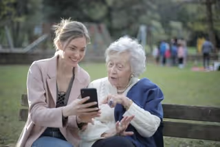 A young caregiver shows a smartphone to an elderly woman on a park bench outdoors.