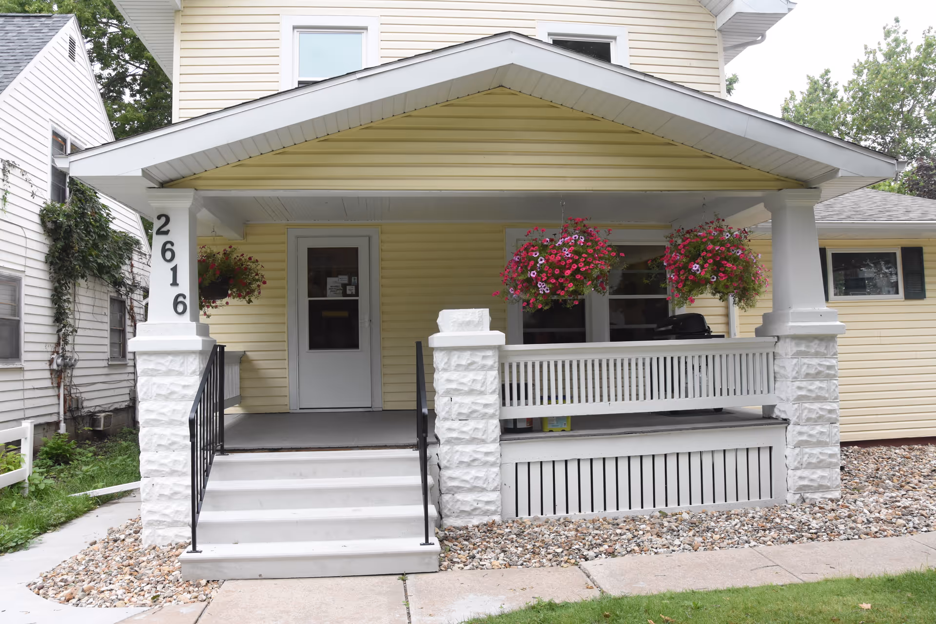 Front porch of a yellow house with hanging flower baskets and steps.