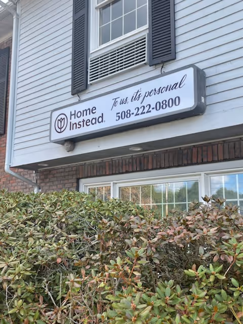 Residential senior care facility sign reading Home Instead on a light-siding building with shrubs.