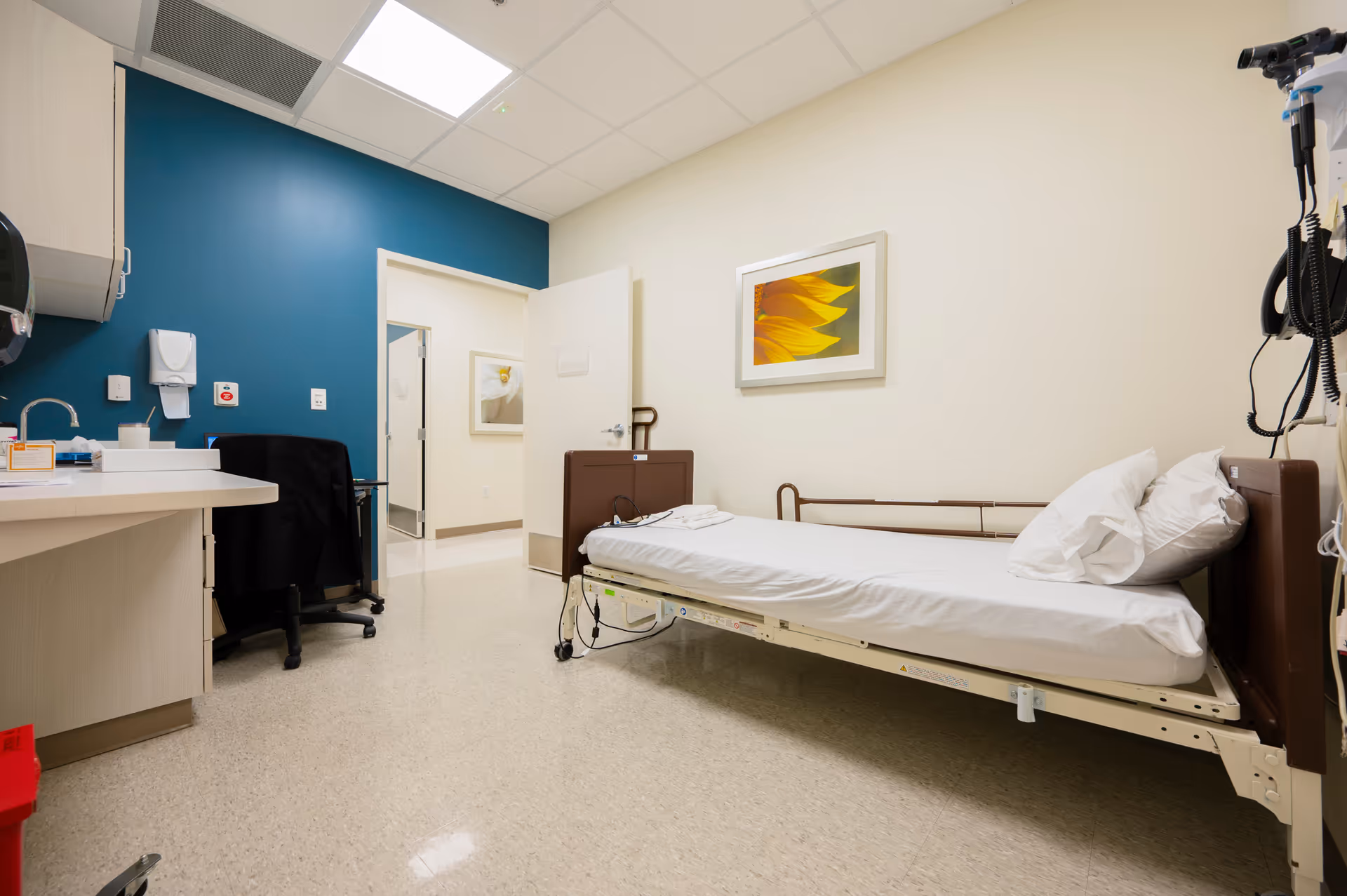 A hospital-style patient room with a single bed, blue accent wall, desk, and medical equipment.