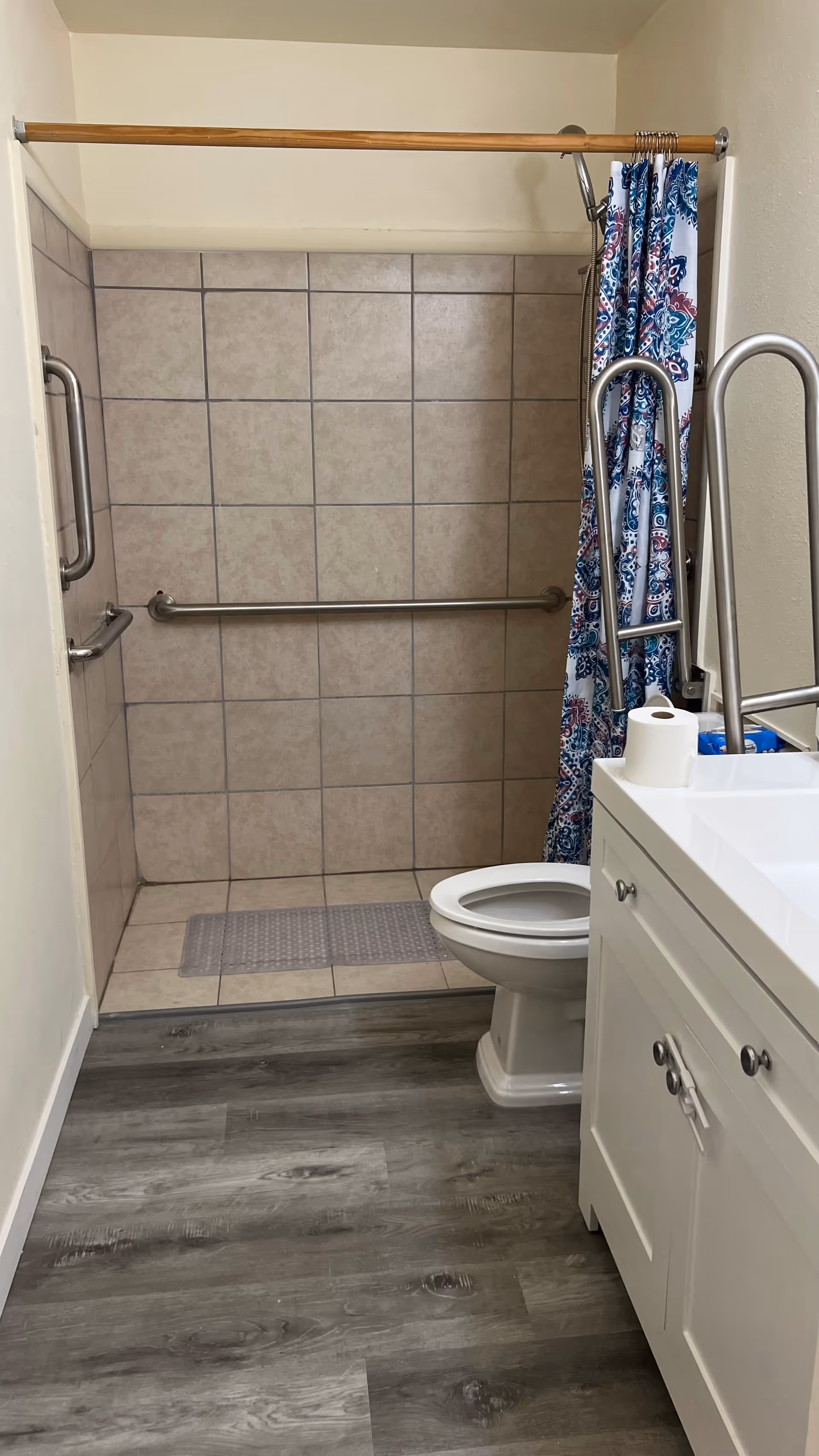 Small bathroom with tiled shower, safety grab bars, toilet, and white vanity.