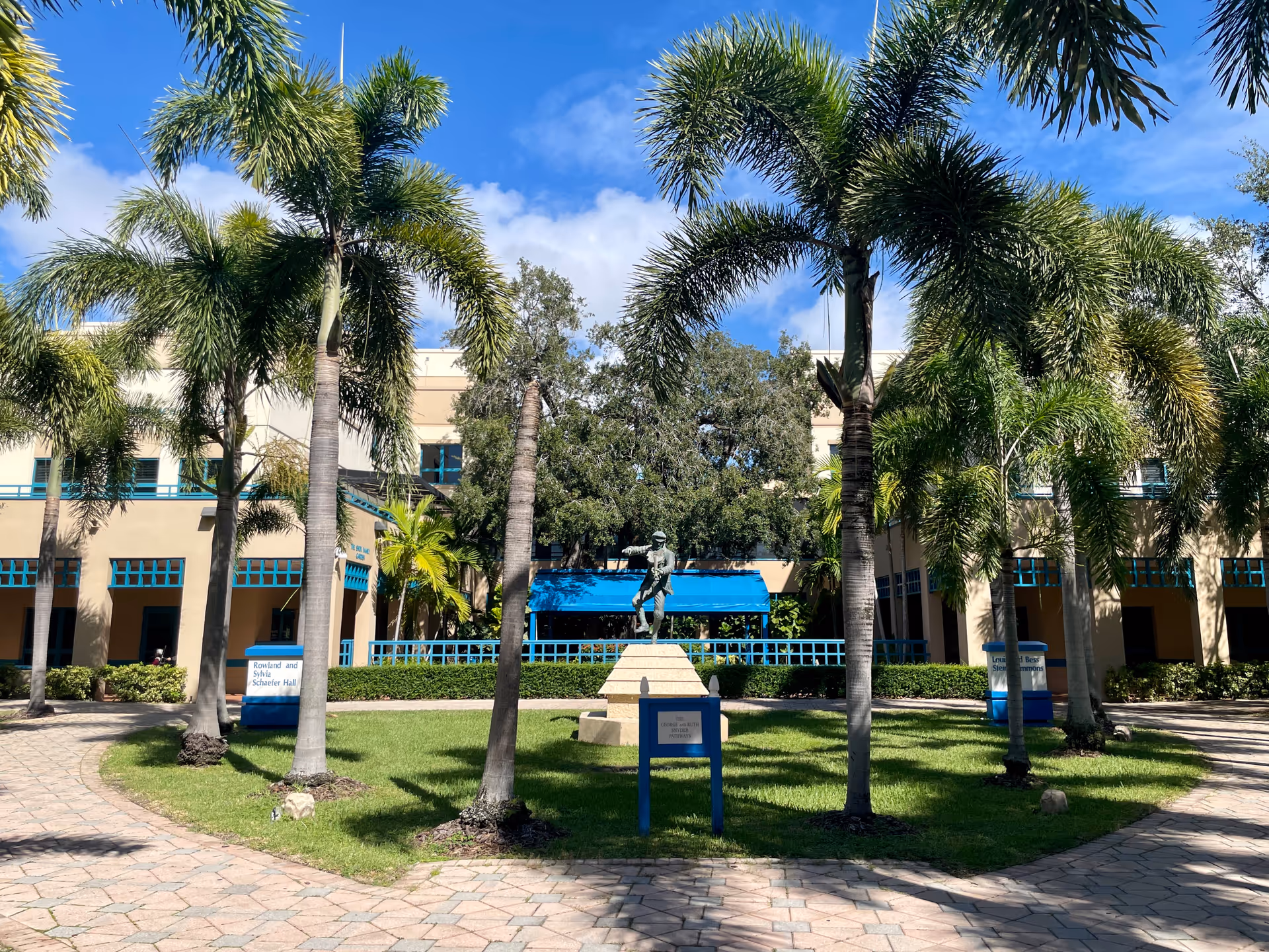 Sunny courtyard with palm trees, a statue, and a beige building in the background.