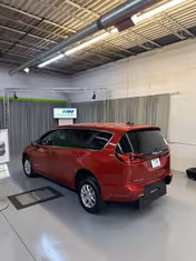 Red minivan parked indoors in a showroom with industrial ceiling and gray curtains.