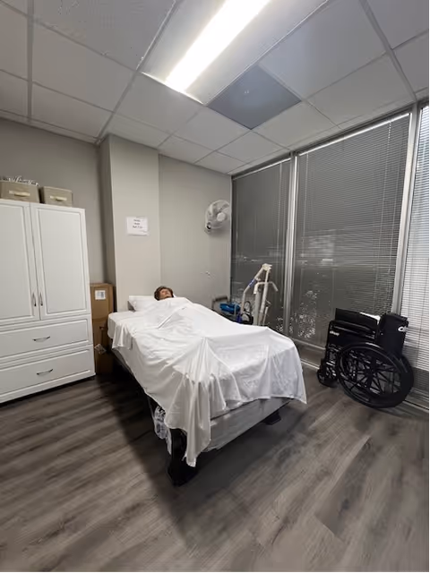 Patient lies in a hospital-style bed in a furnished care room with a wheelchair.
