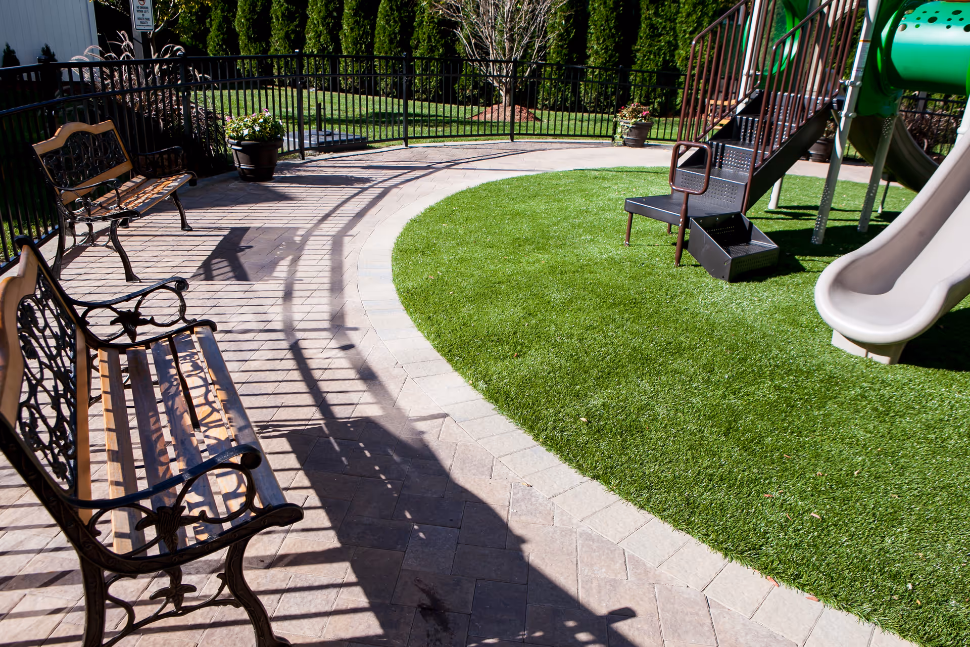 Outdoor playground area with curved walkway, benches, and green grass near a fence.