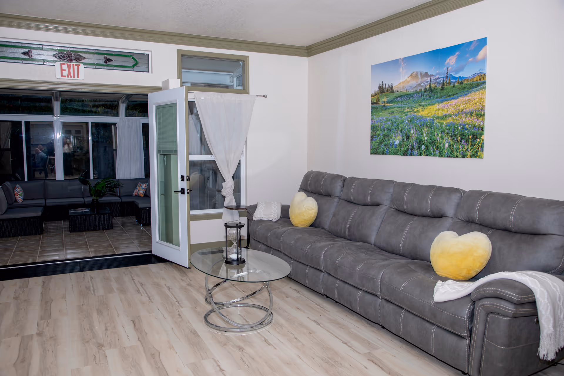 Cozy common living room with gray sectional sofa, heart cushions, glass coffee table, and exit door.