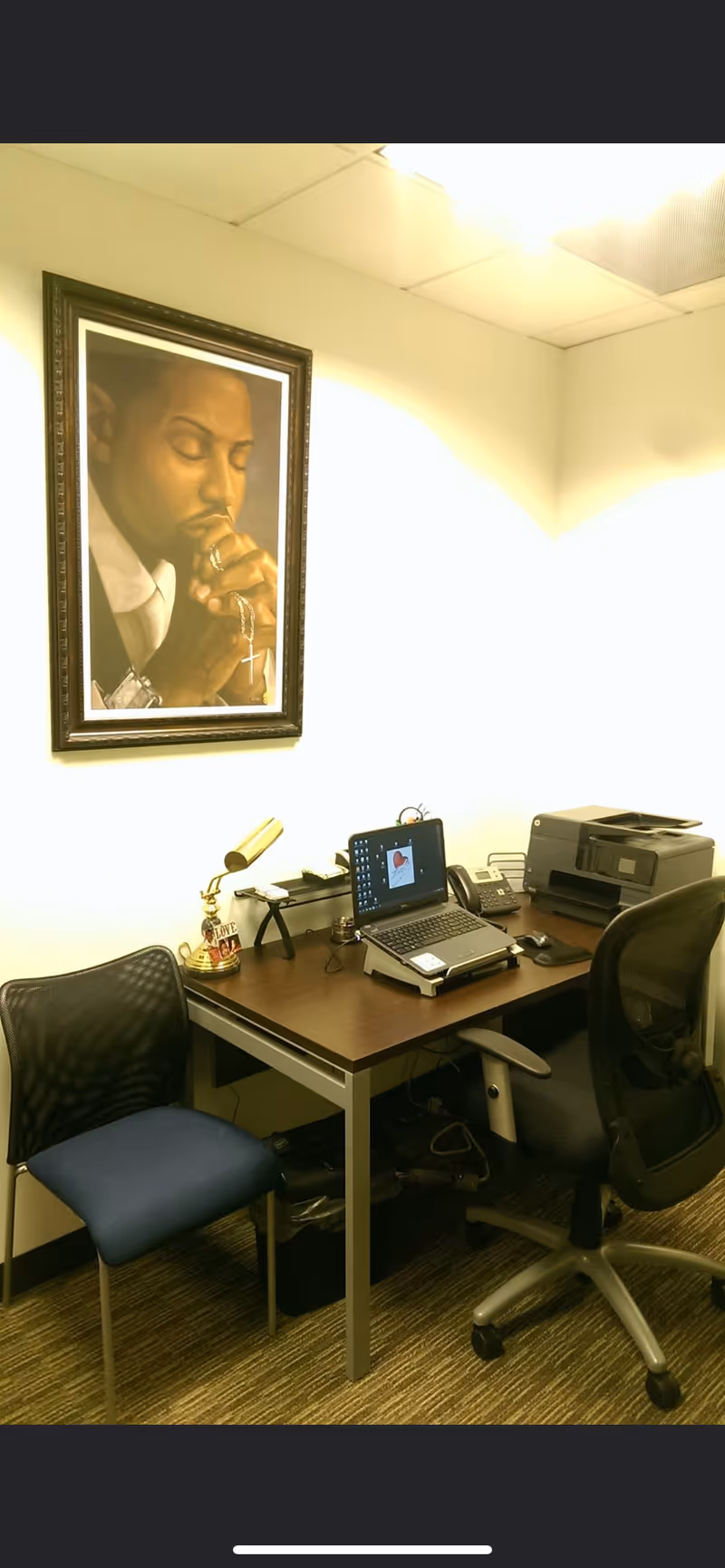 Interior office area with desk, laptop, printer, lamp, and framed portrait on wall.