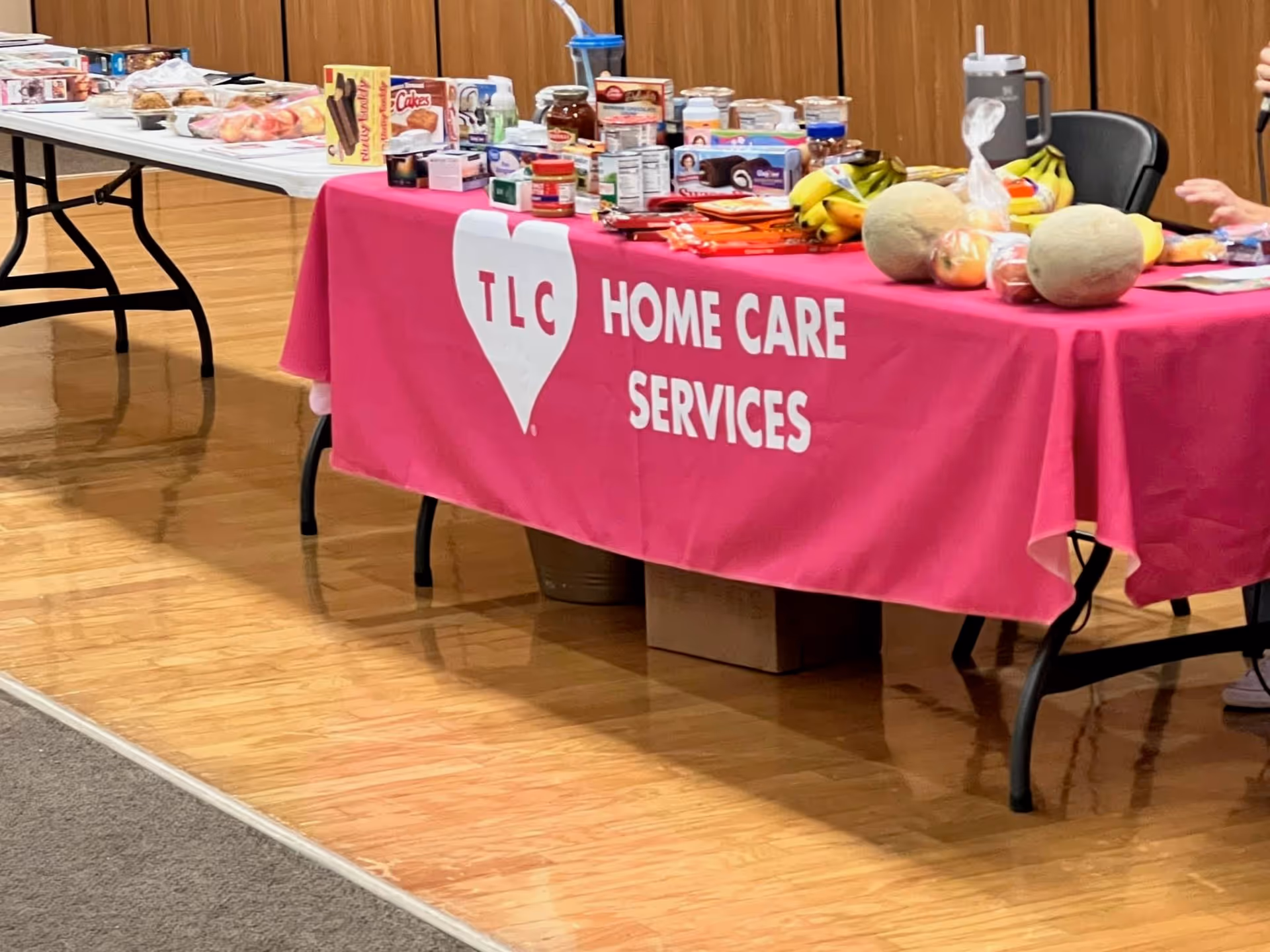 Indoor hall with a pink TLC Home Care Services table displaying assorted food and items.