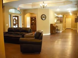 Wide interior lounge with leather sofas, a clock, chandelier, and kitchen area in background.