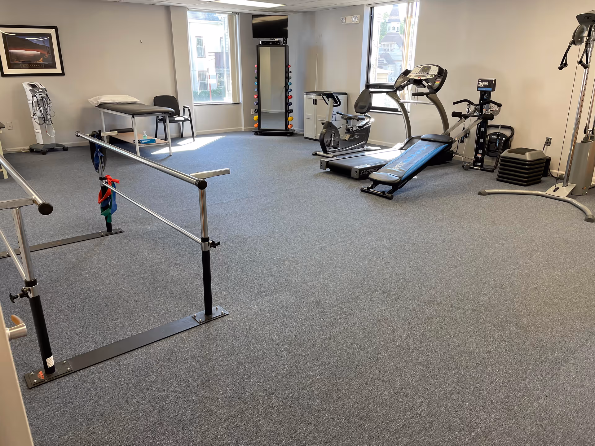 A fitness and therapy room with treadmills, bikes, parallel bars, and a treatment table.