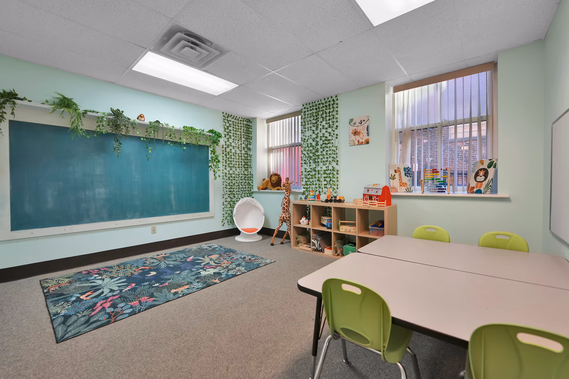 Bright classroom-like playroom with chalkboard, shelves, plants, and child-sized tables and chairs.