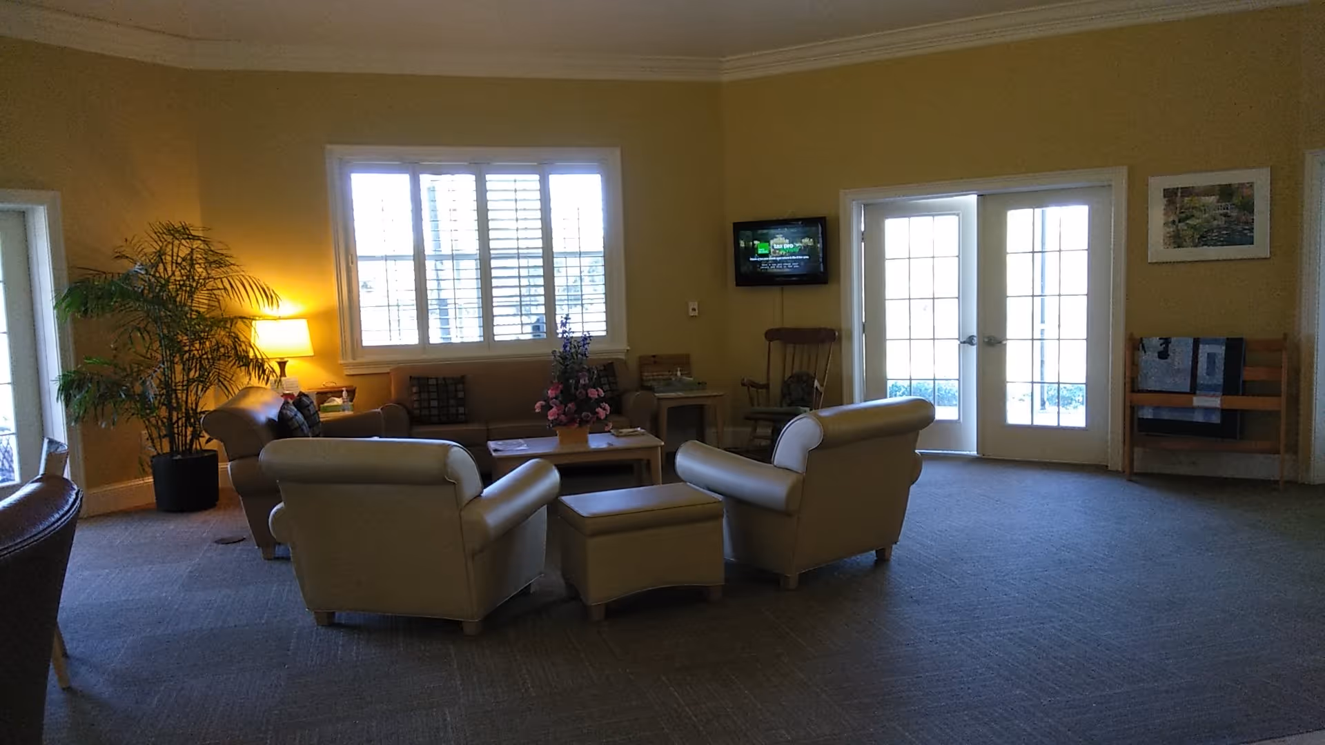 A spacious senior living lounge with beige chairs, a coffee table, plants, and a wall TV.